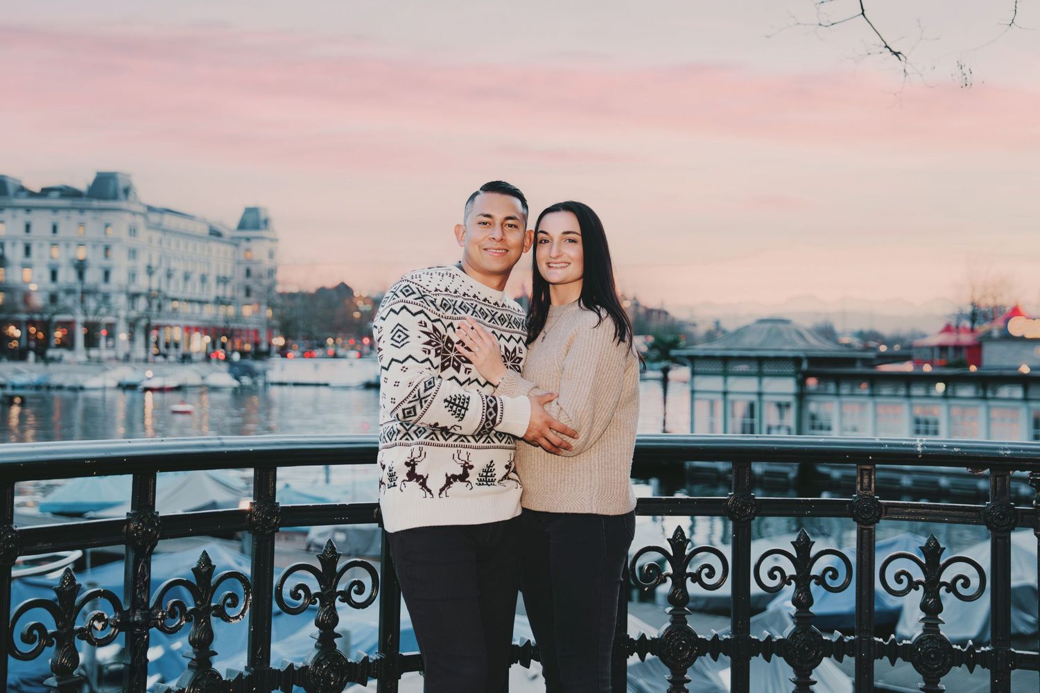 Smiling couple in matching holiday sweaters posing by ornate railing at Zurich waterfront.