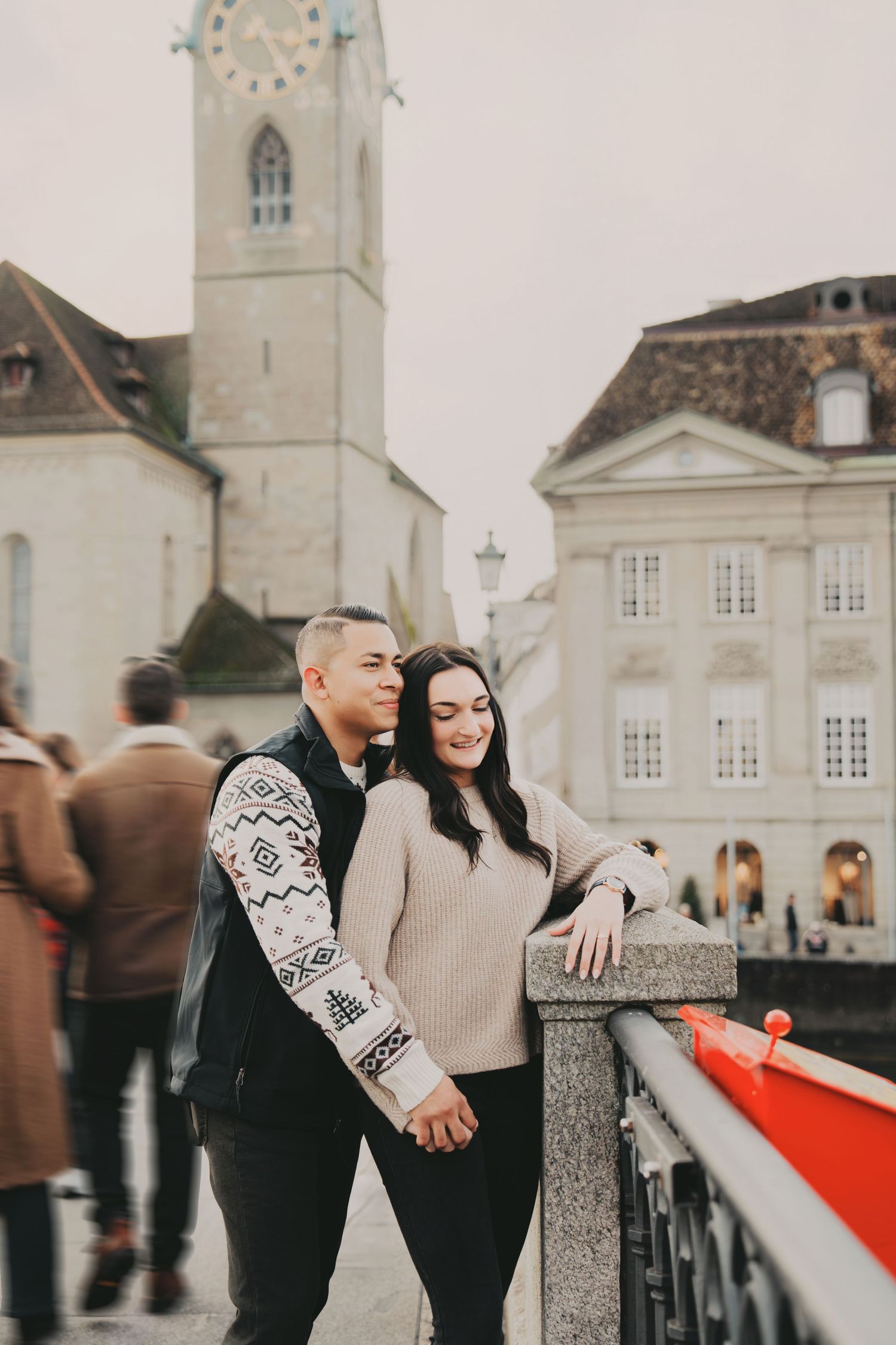 Couple holding hands on a busy Zurich bridge with a tall historic church tower and city buildings behind.