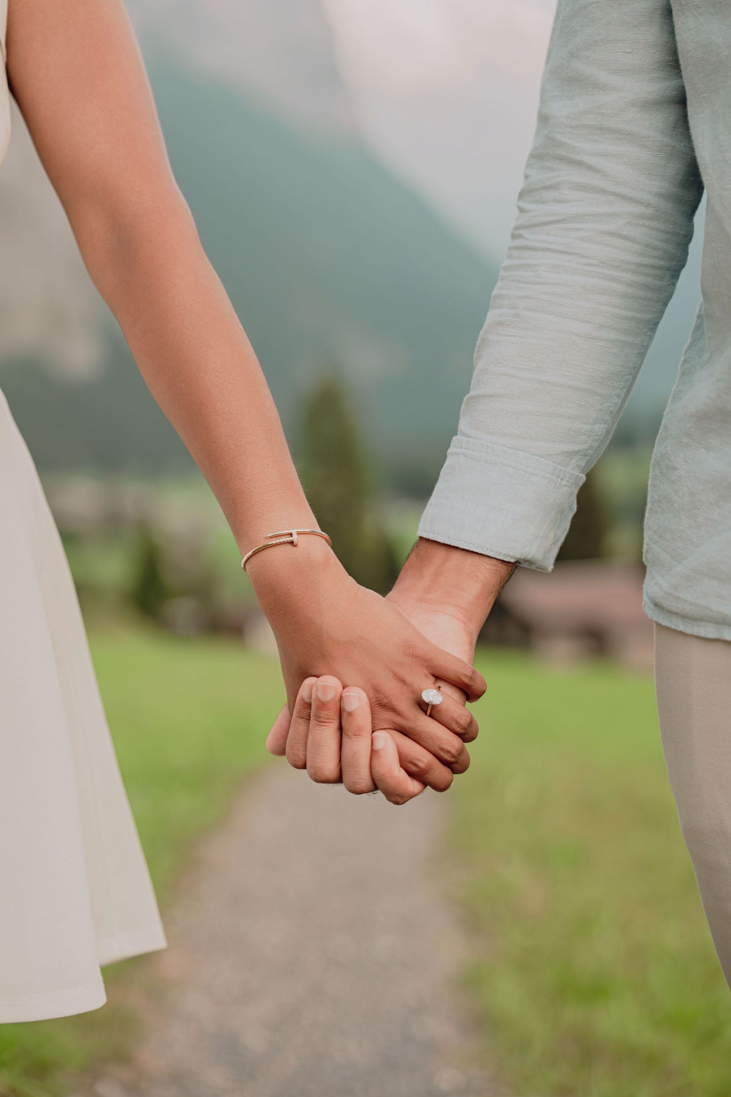 Close-up of an engaged couple holding hands on a path, woman wearing diamond ring and gold bracelet.