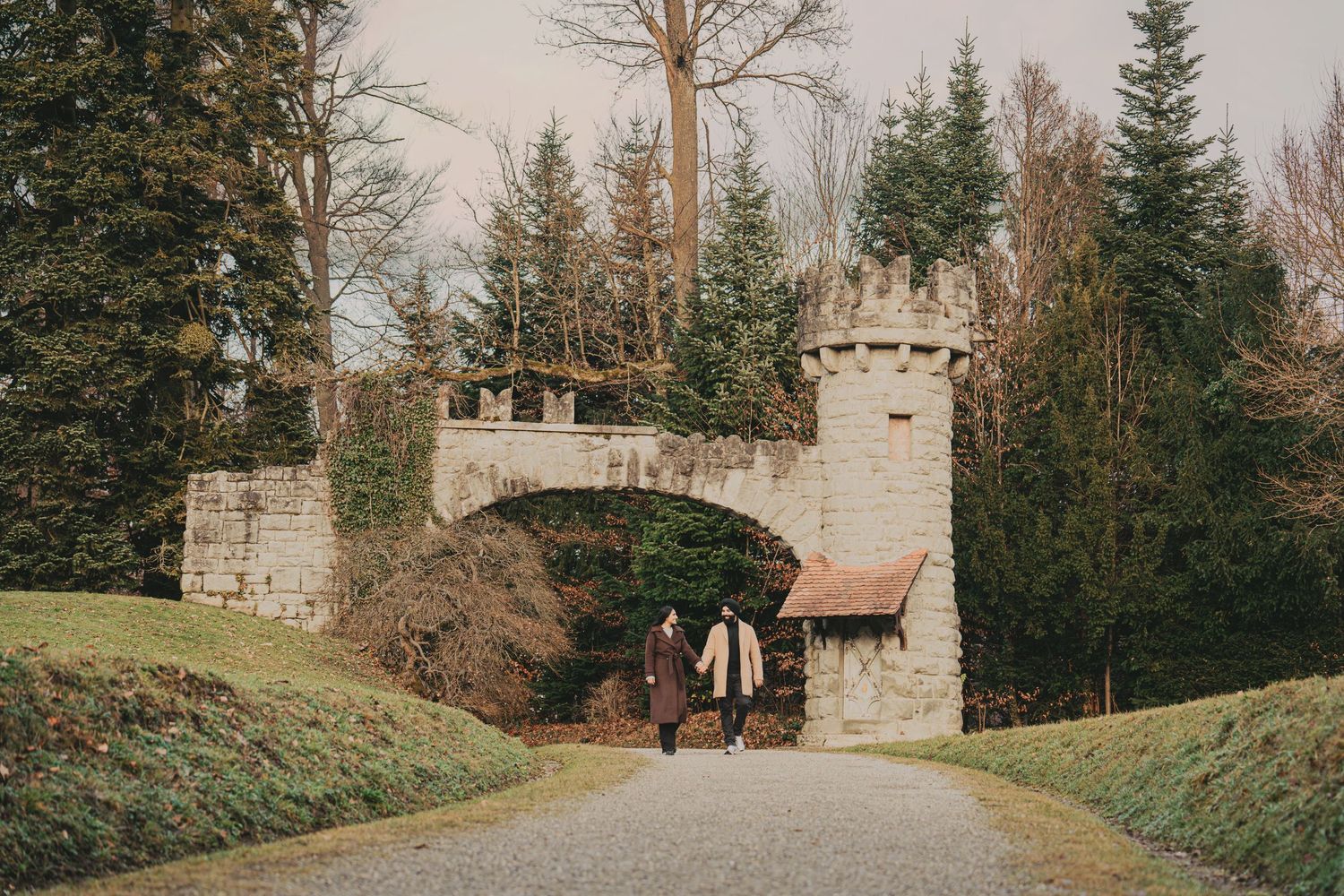 Engaged couple walking through a stone castle gate archway surrounded by tall evergreen trees.