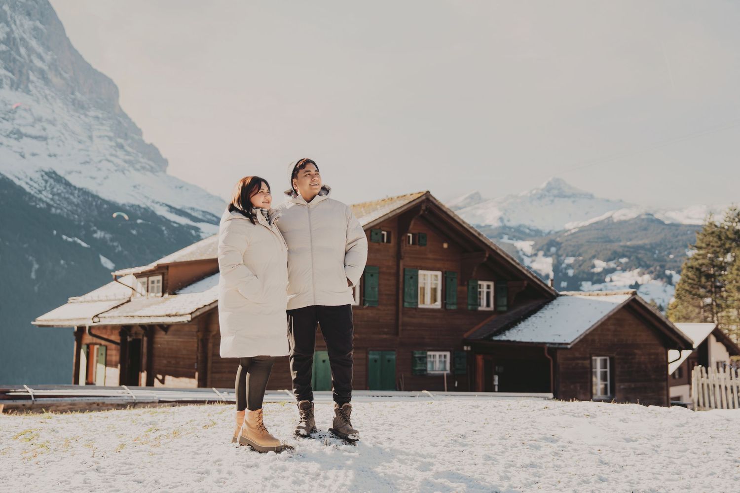 Couple standing together in white puffer coats on snow with traditional Swiss chalet behind them.