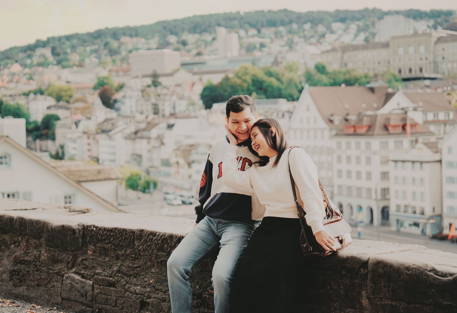 Romantic couple sitting close together on stone wall with Zurich cityscape in background.