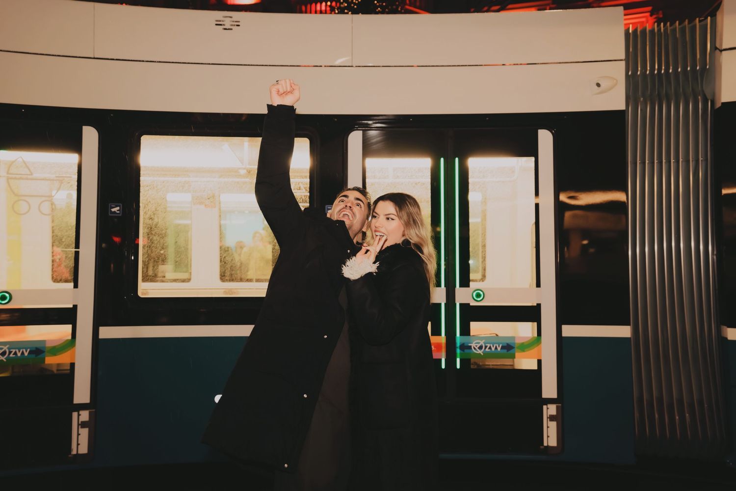 Playful couple posing together in front of a metro train at night.