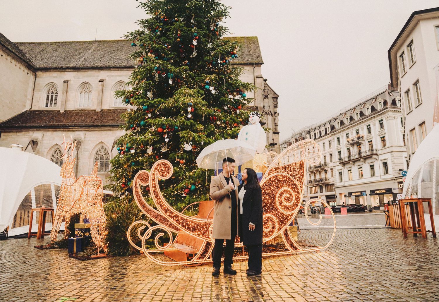 Couple kissing under umbrella by Christmas tree and festive sleigh decor.