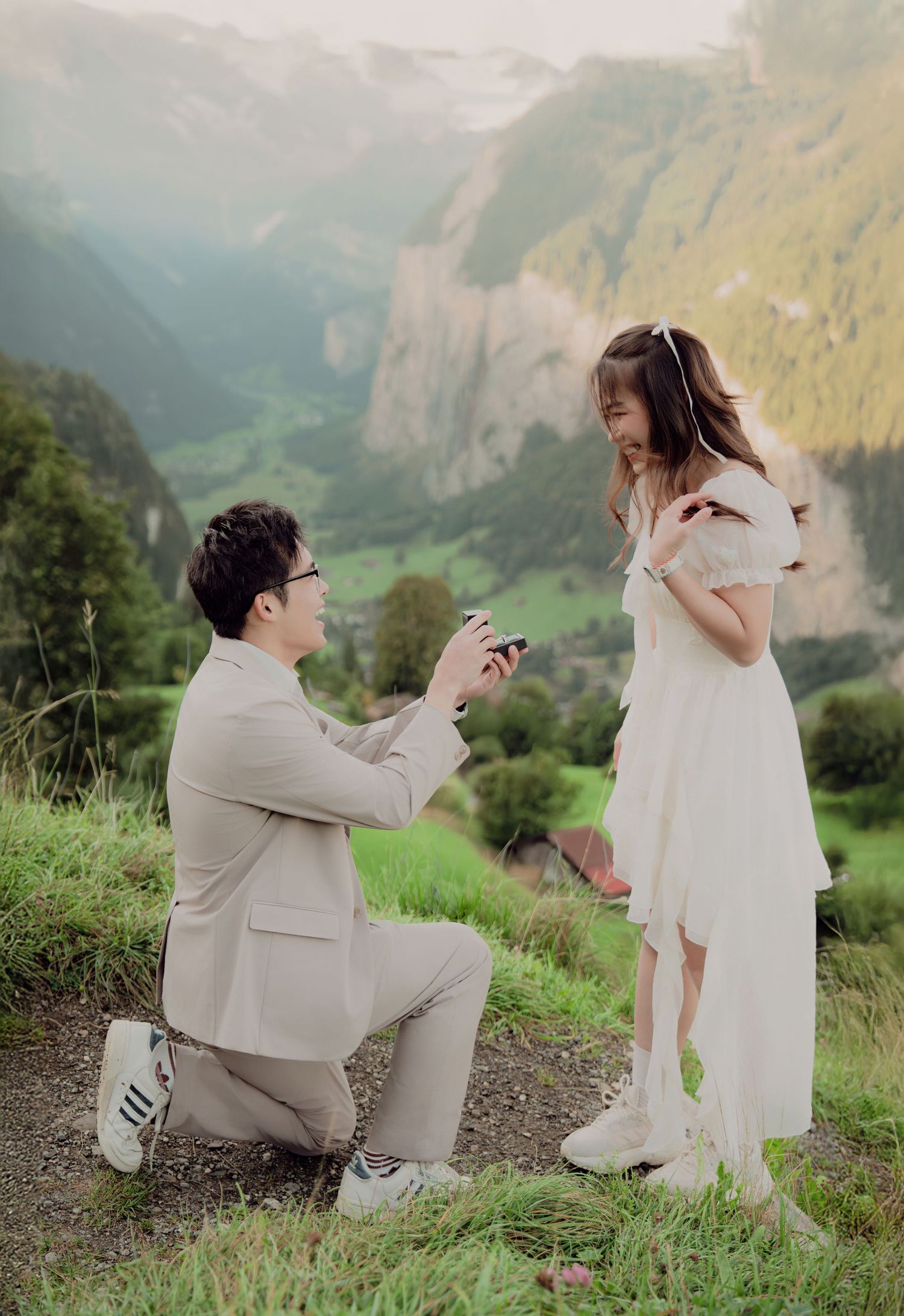Man kneeling on one knee proposing to woman in white dress with stunning Swiss Alps valley backdrop.