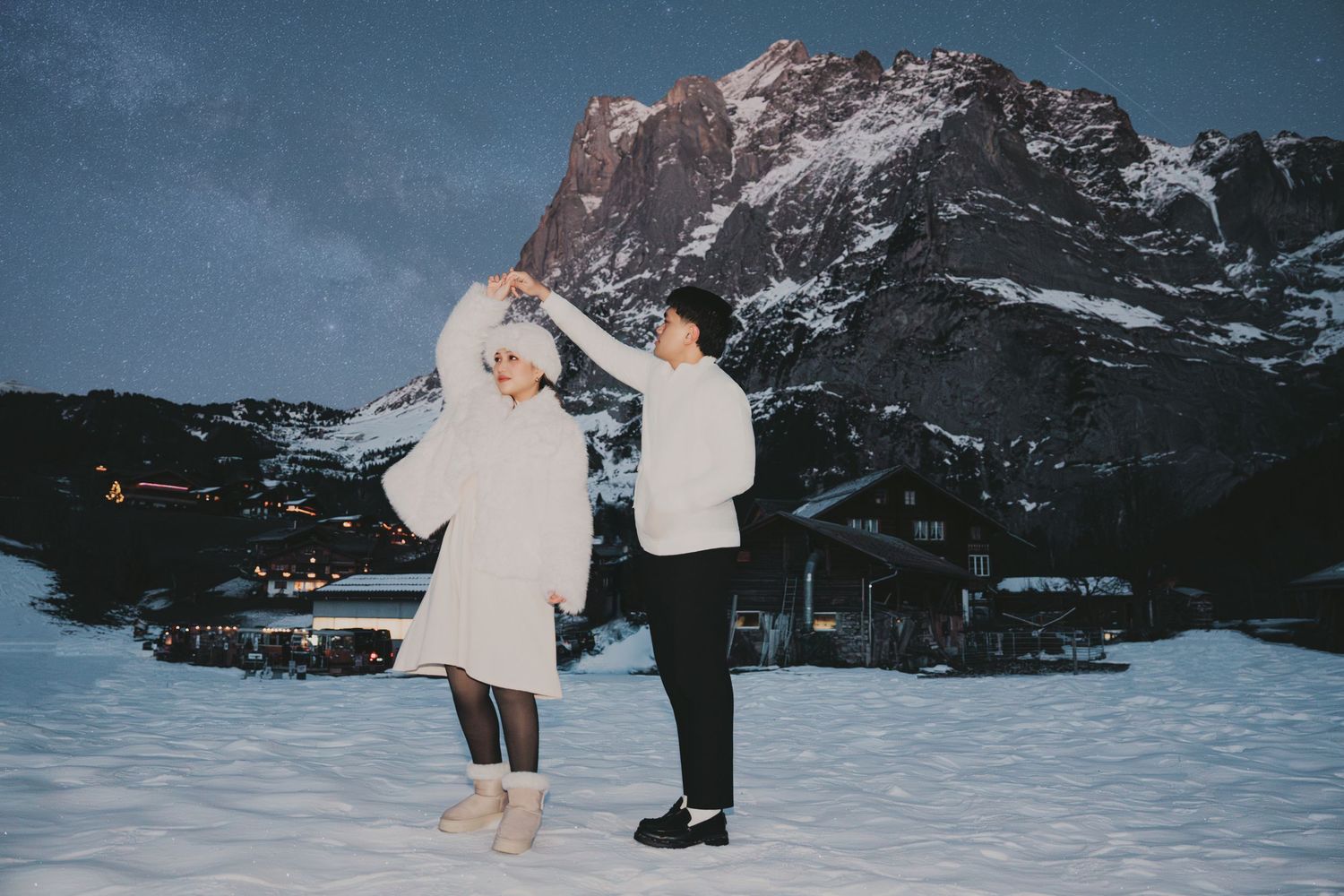 Couple spinning and dancing on snow at night with illuminated alpine village and mountain backdrop.