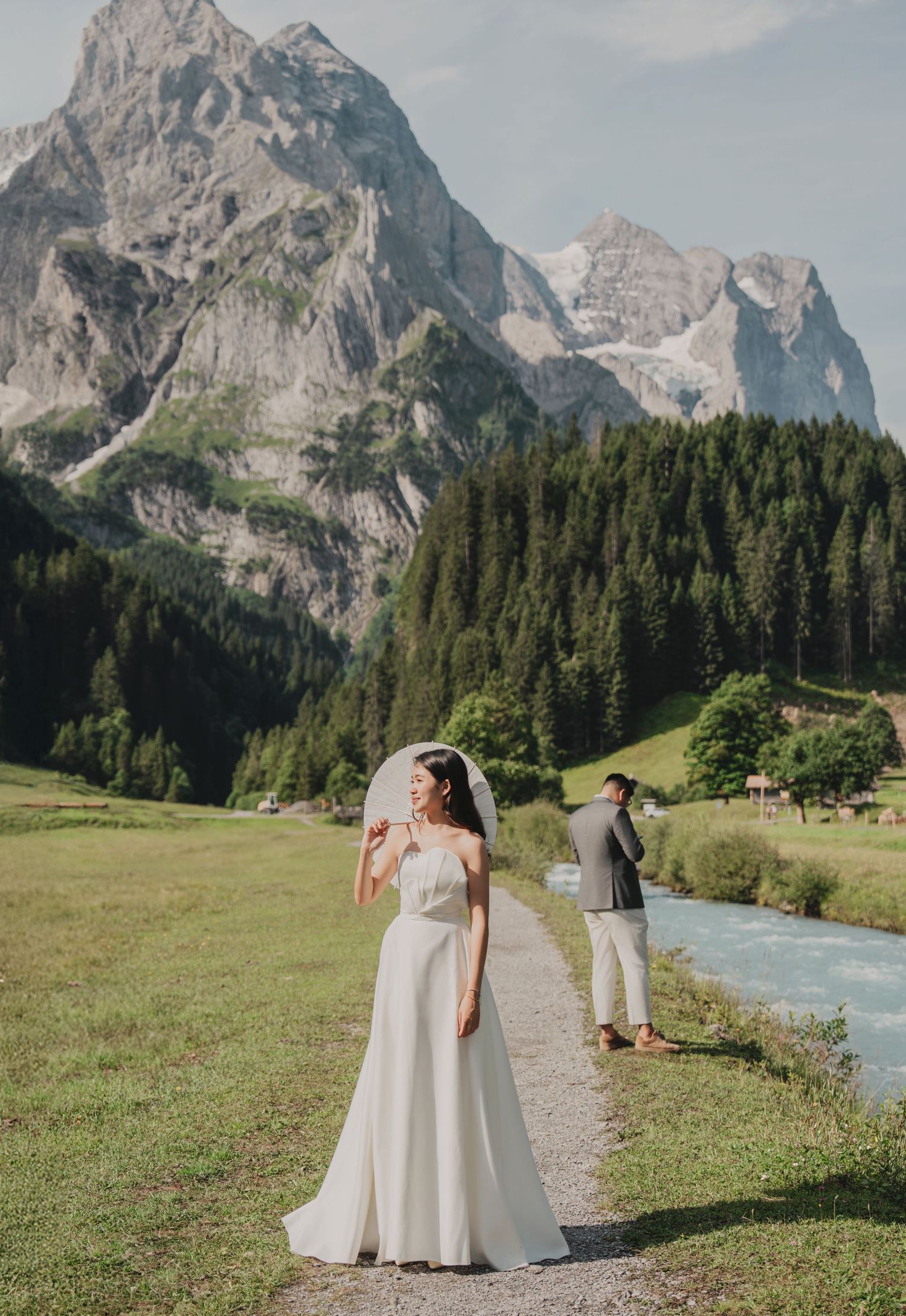 Bride in white gown with veil stands on alpine path as groom looks toward Swiss mountains.