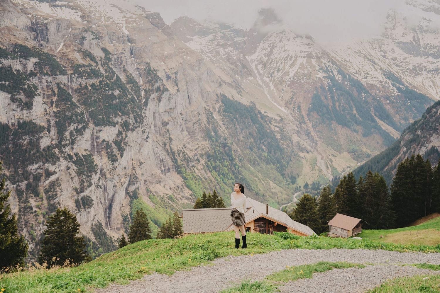 Lone woman stands on gravel alpine path with dramatic rocky mountain cliffs and wooden chalets below.