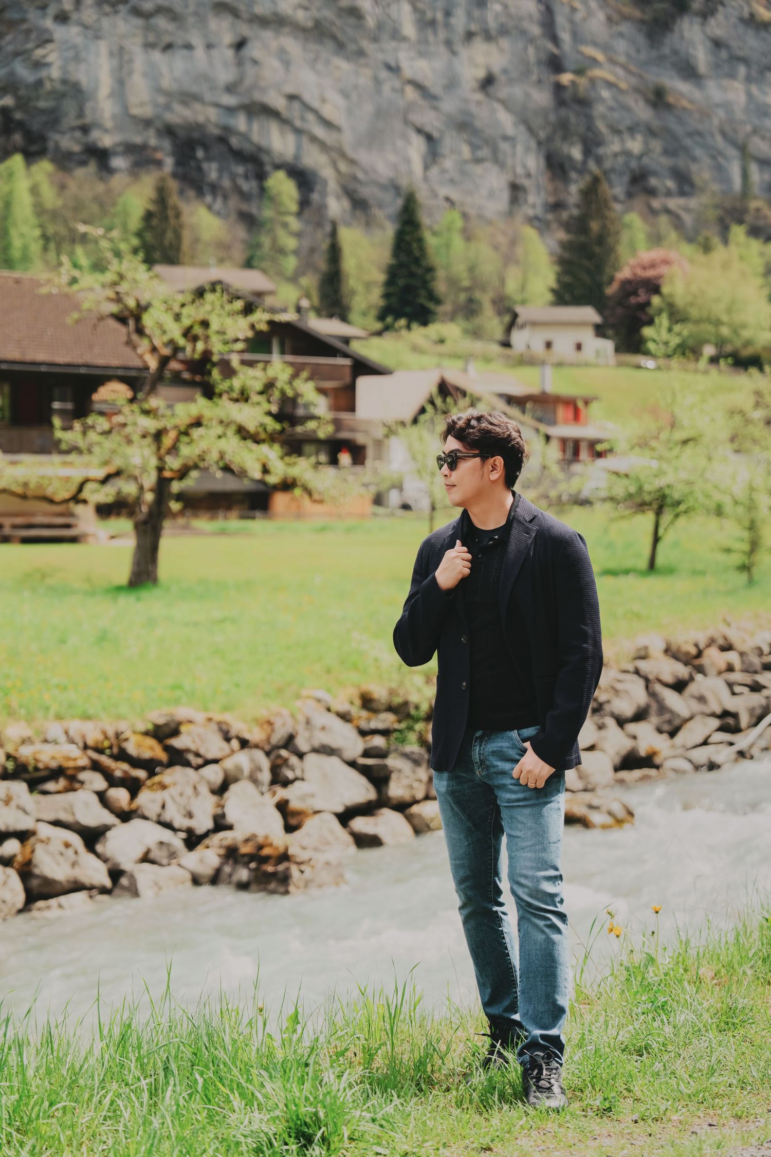 Man in dark blazer and jeans standing by rocky river in green Swiss mountain landscape.
