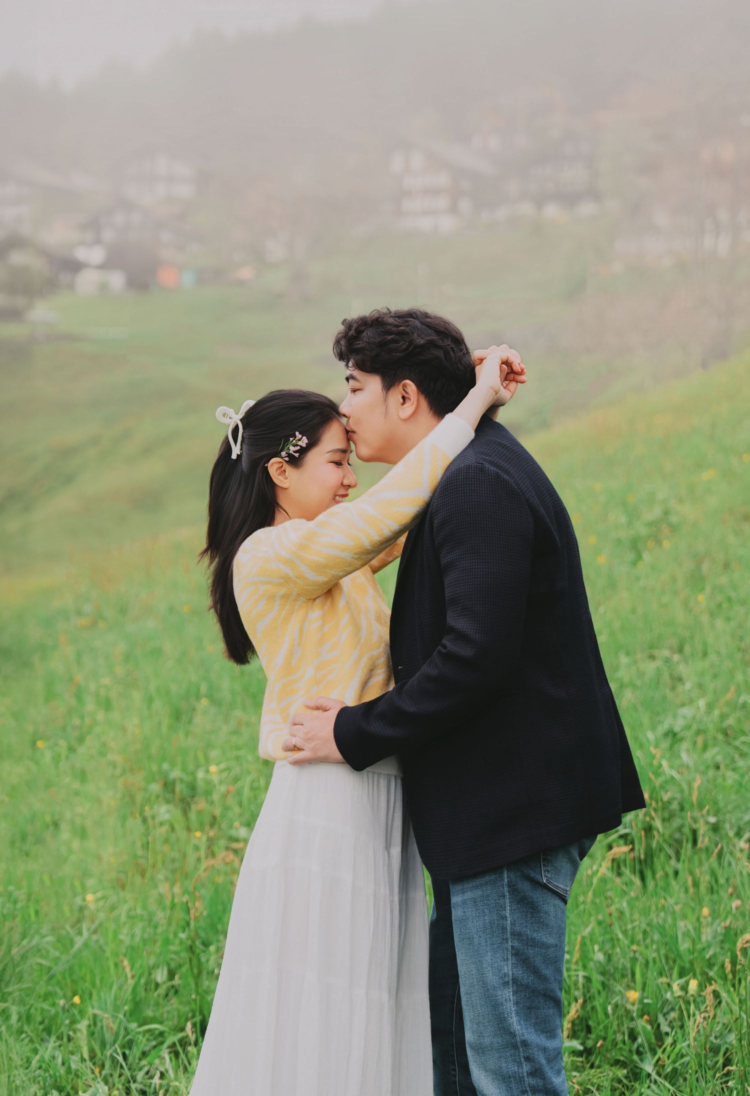 Couple sharing a tender embrace and kiss on a misty green hillside with village in background.