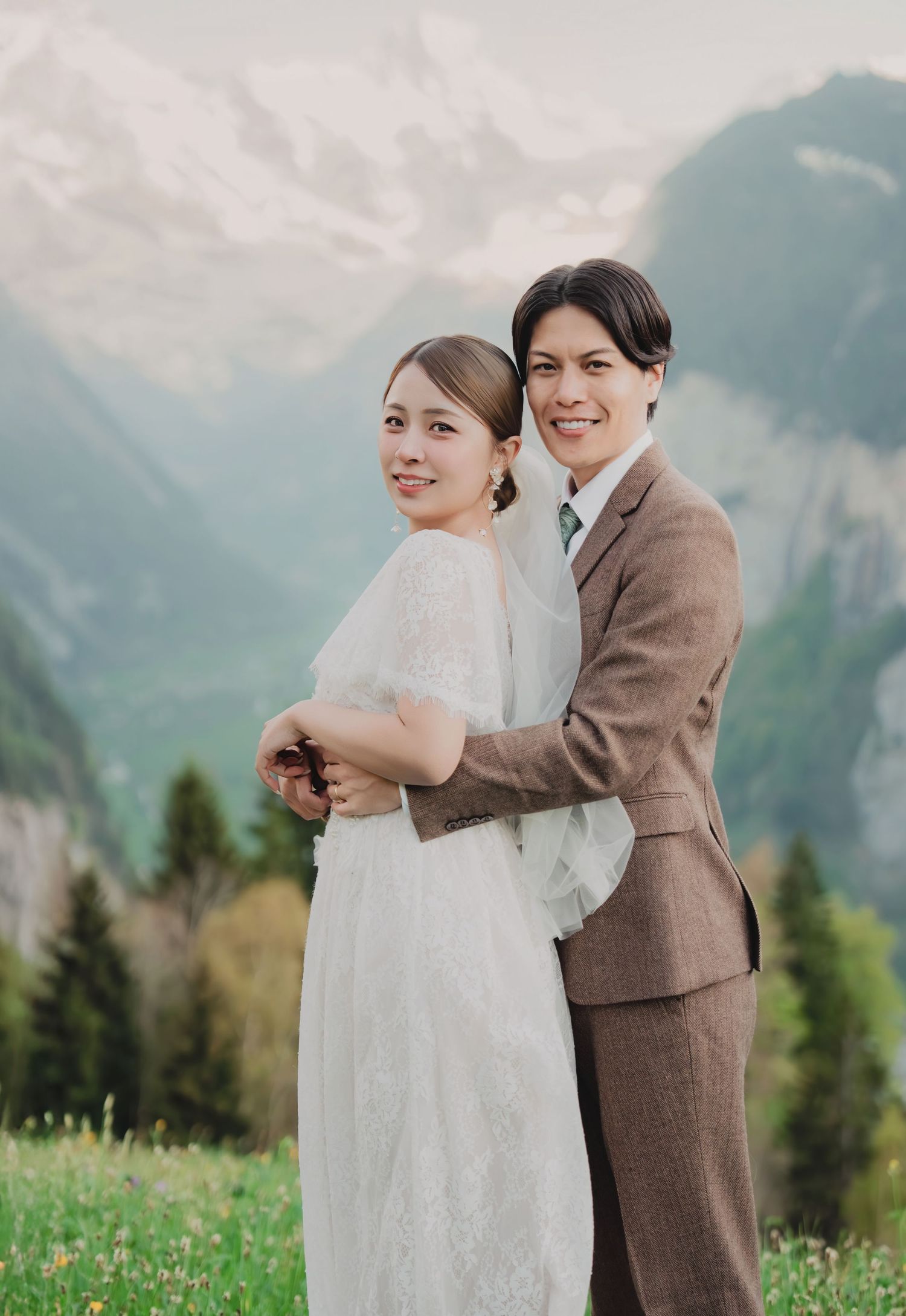 Couple in wedding attire embracing on alpine meadow with snow-capped Swiss mountains backdrop.