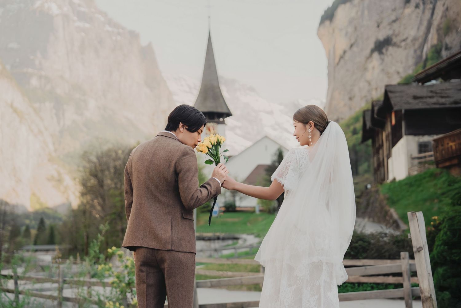 Bride and groom exchanging flowers near a charming Swiss church surrounded by misty alpine scenery.
