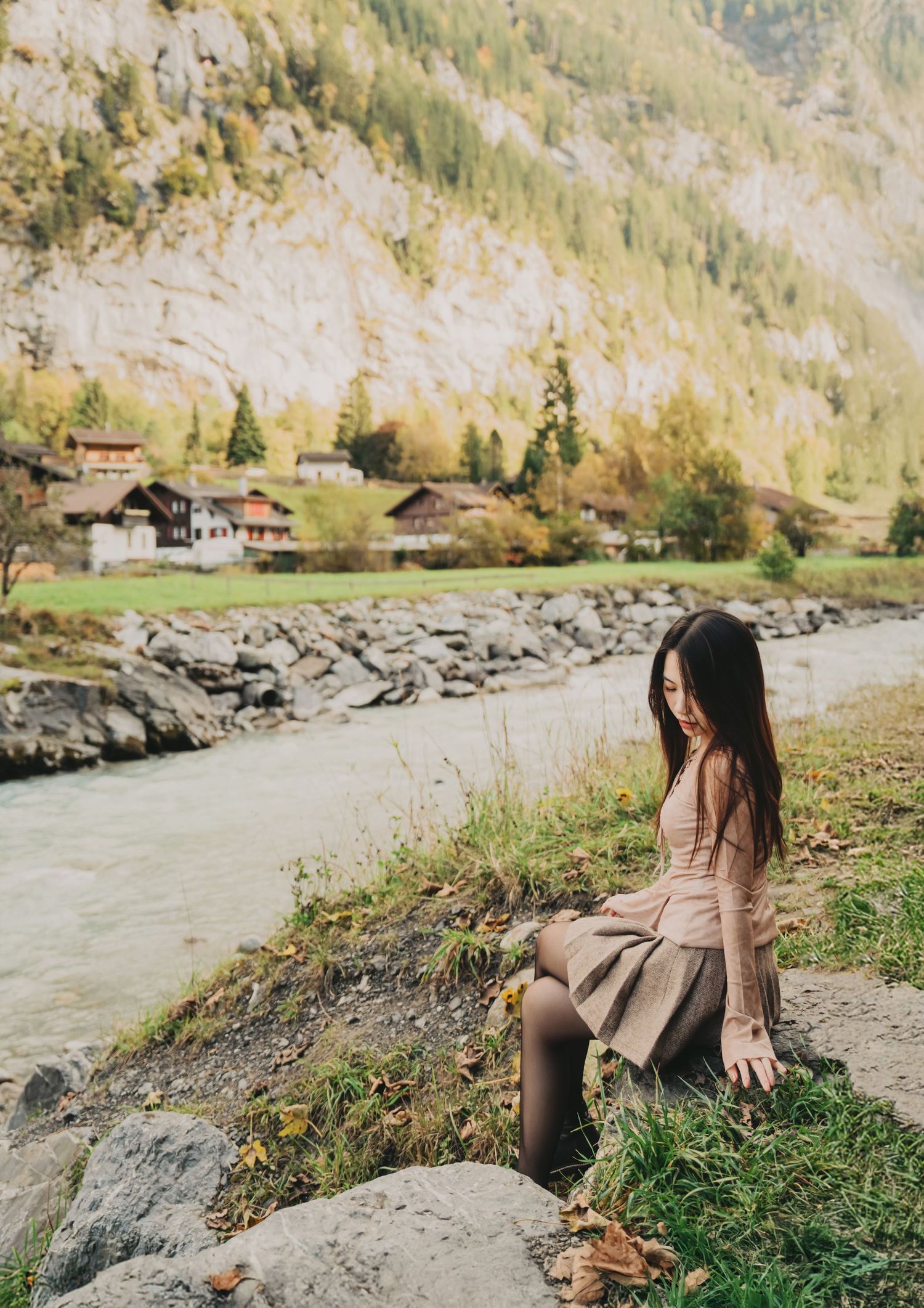 Young woman sitting on rocks beside a serene river in a stunning Swiss alpine landscape.