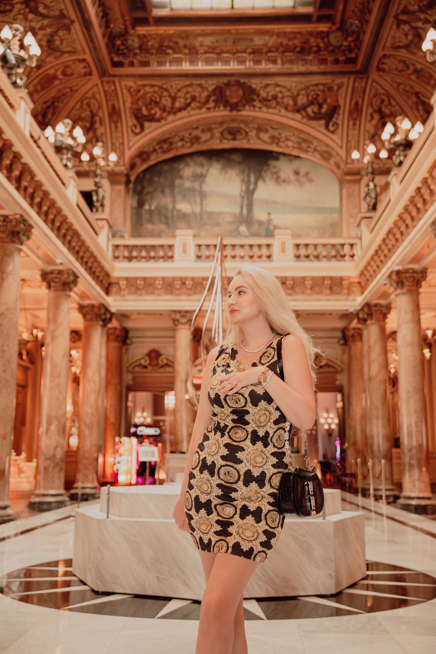 Blonde woman in Versace-style patterned mini dress posing inside an ornate gilded casino hall in Monaco.