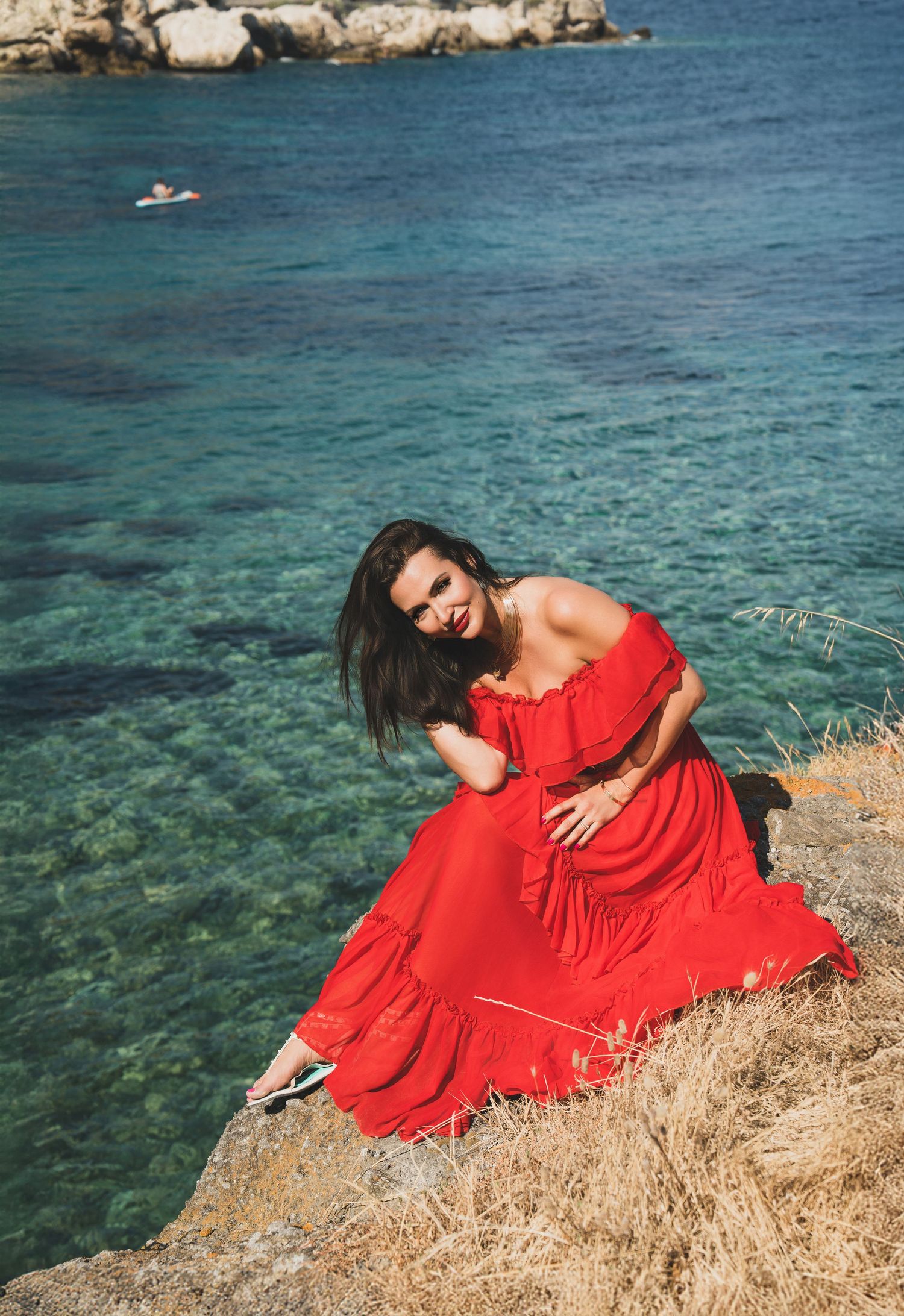 Smiling woman in red off-shoulder dress posing on rocky cliff above sparkling turquoise sea.