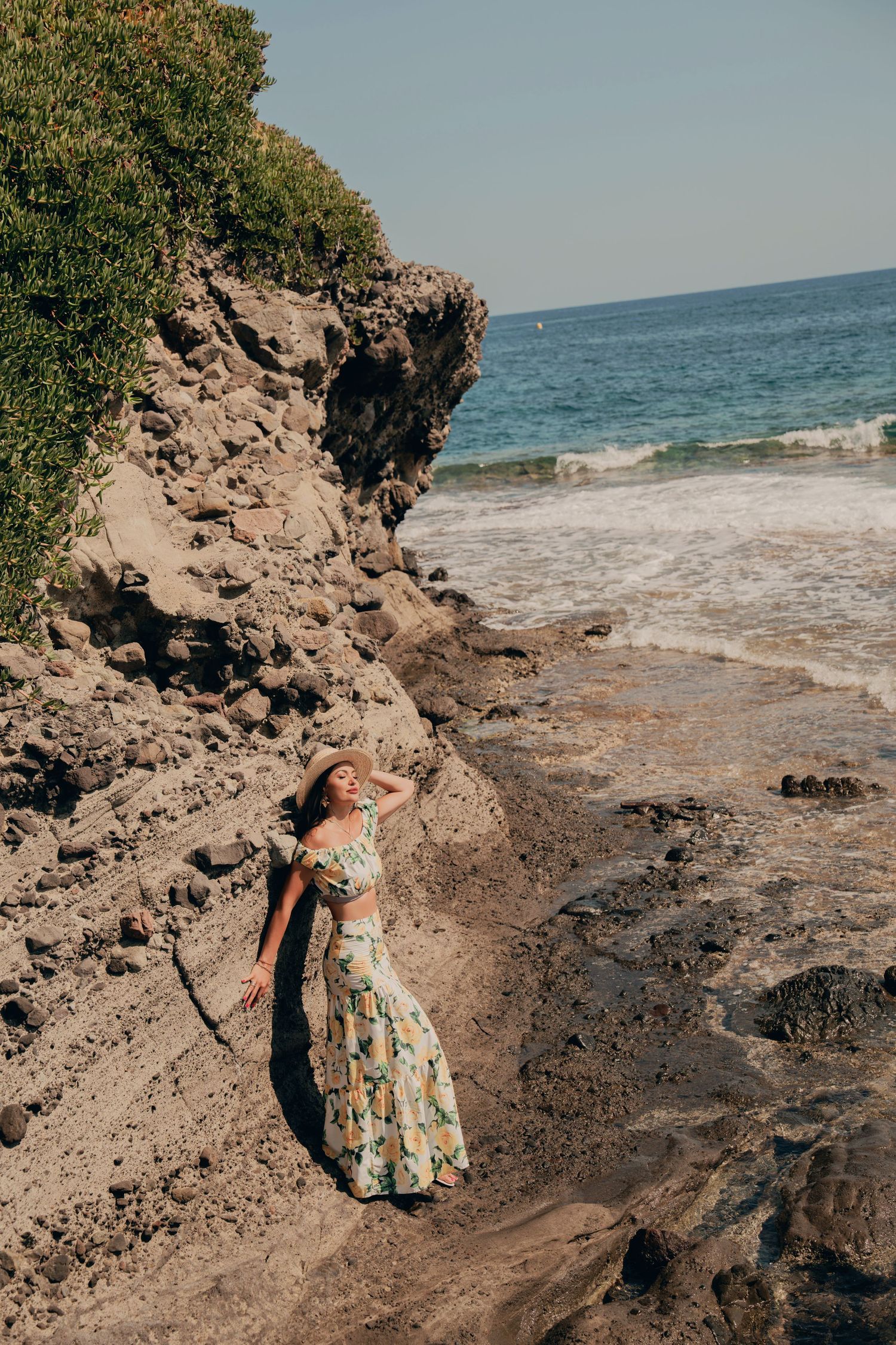 Woman in floral sundress leaning against rocky beach cliffs with turquoise ocean backdrop.