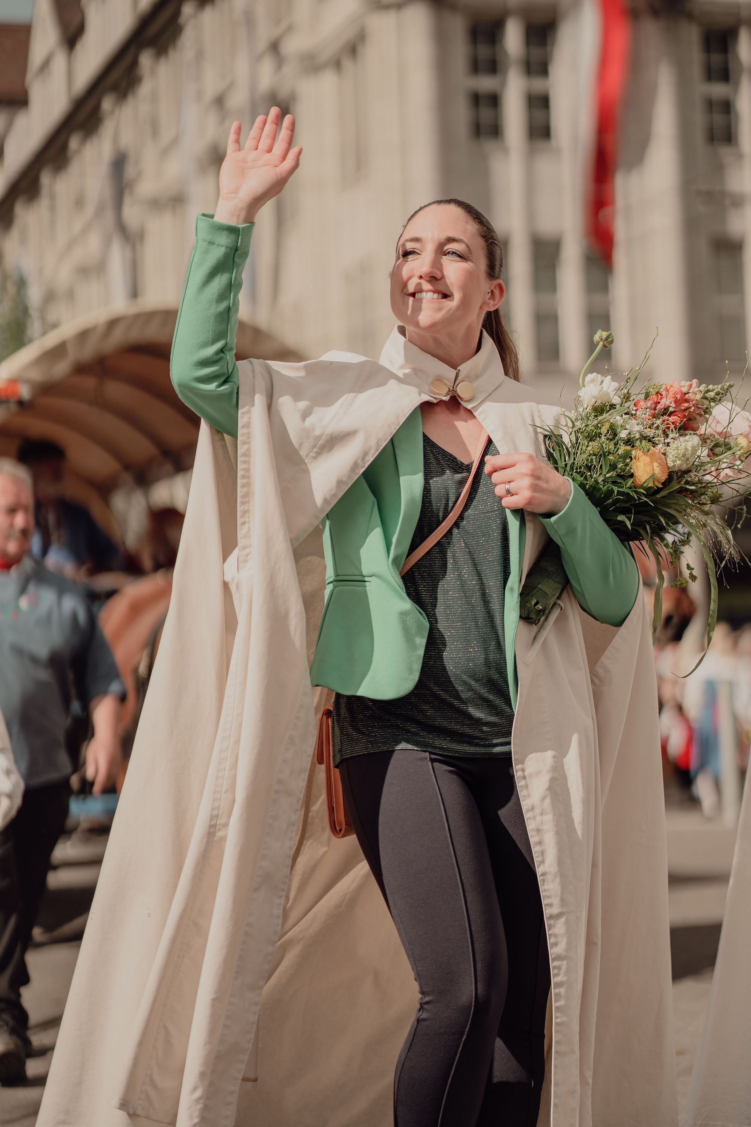 Cheerful woman in white cape and green jacket waves to the crowd while holding flowers at a parade.
