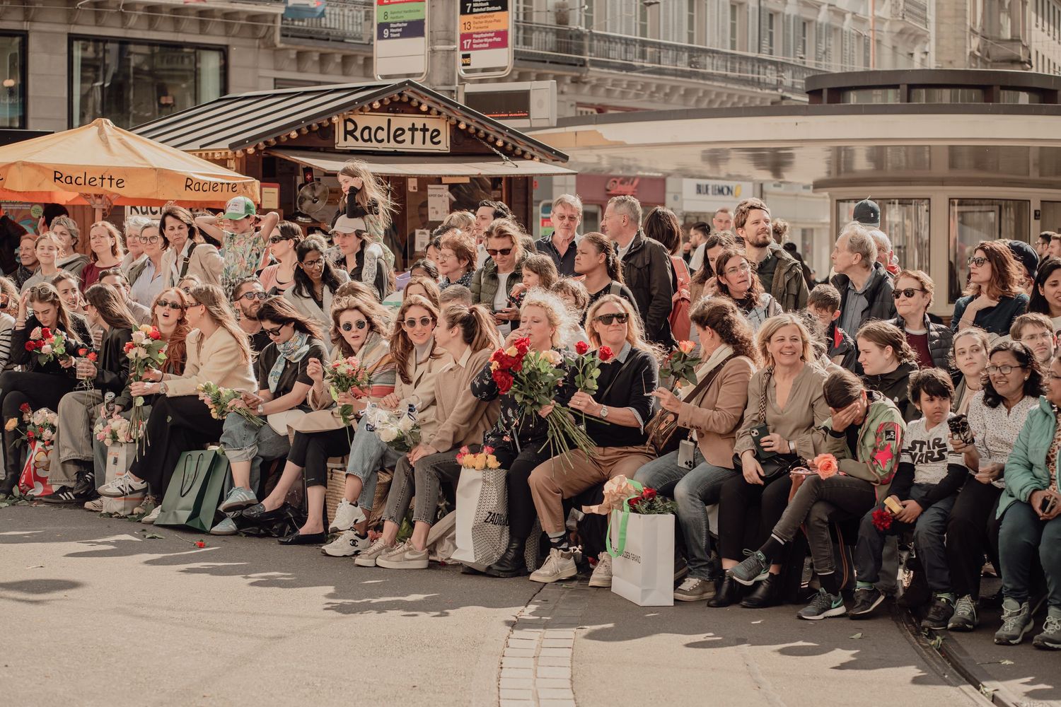 Smiling spectators holding flowers sit closely together watching a traditional cultural street parade.