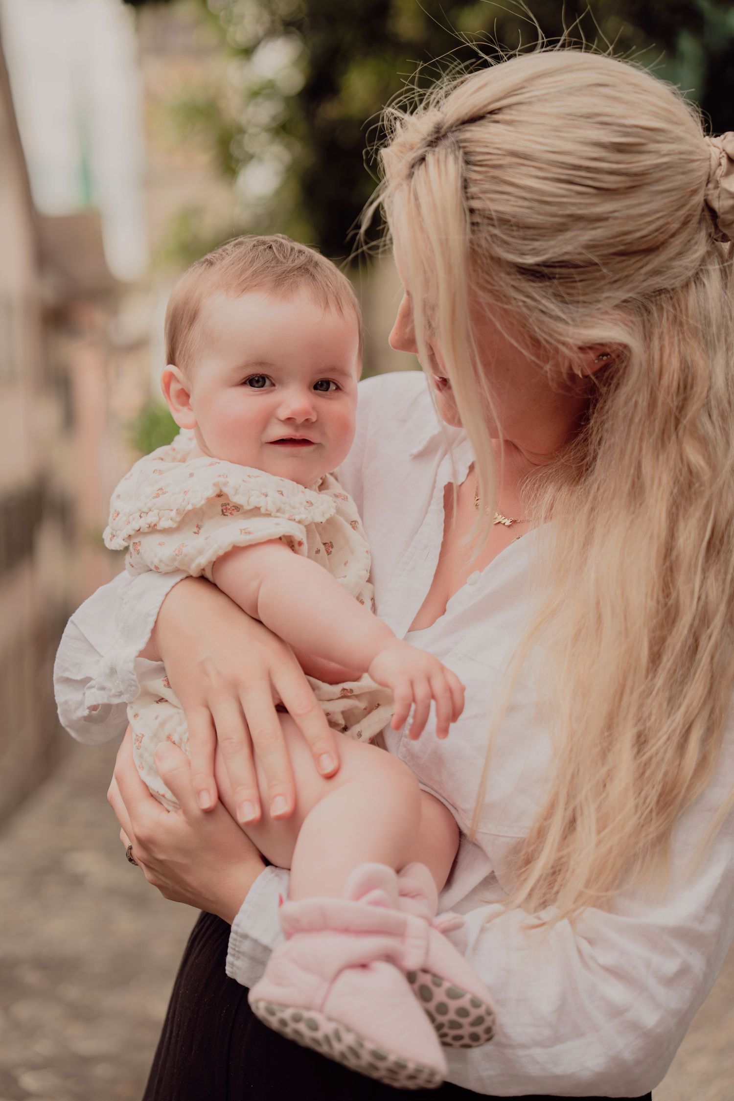 Mother with long blonde hair cradling baby in floral dress on European street.