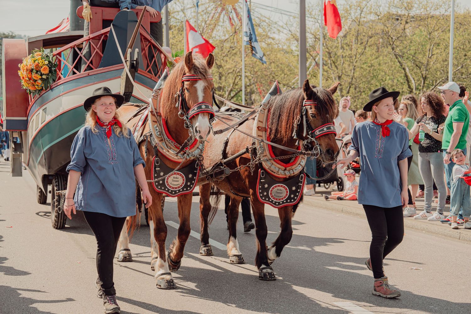 Two handlers lead decorated horses pulling a traditional carriage in a festive parade.
