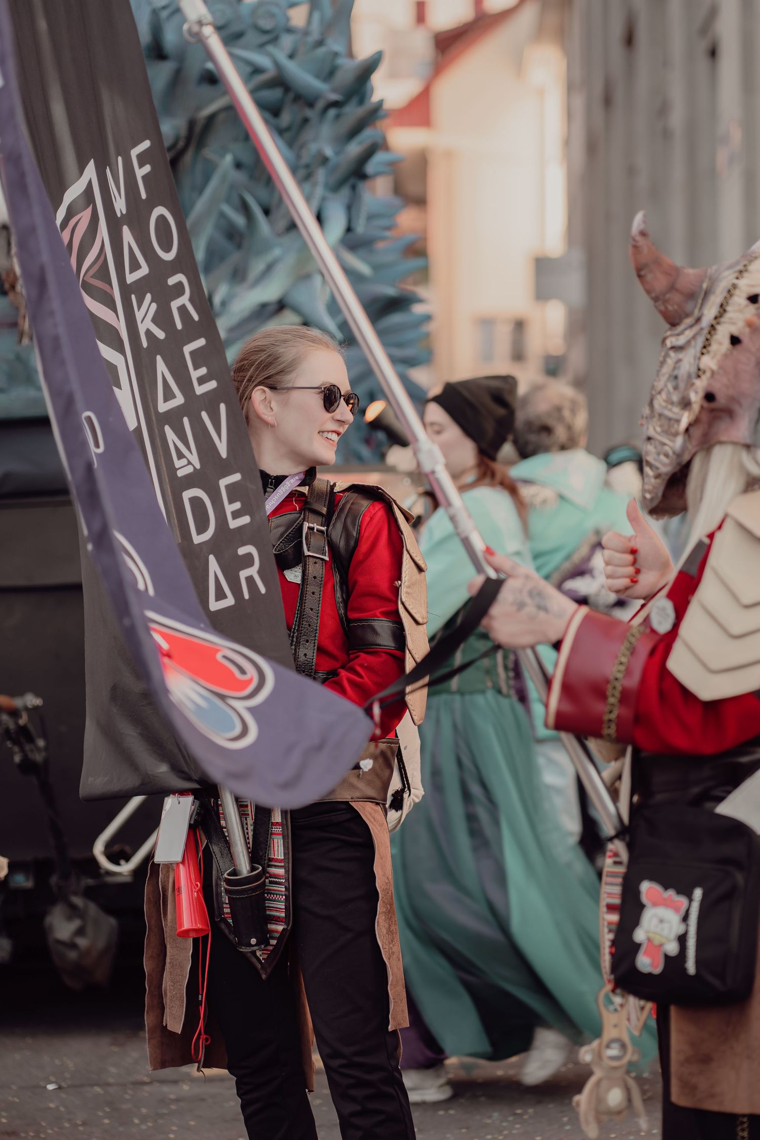 Smiling woman in red jacket holding a flag with runic symbols at an outdoor cosplay festival event.