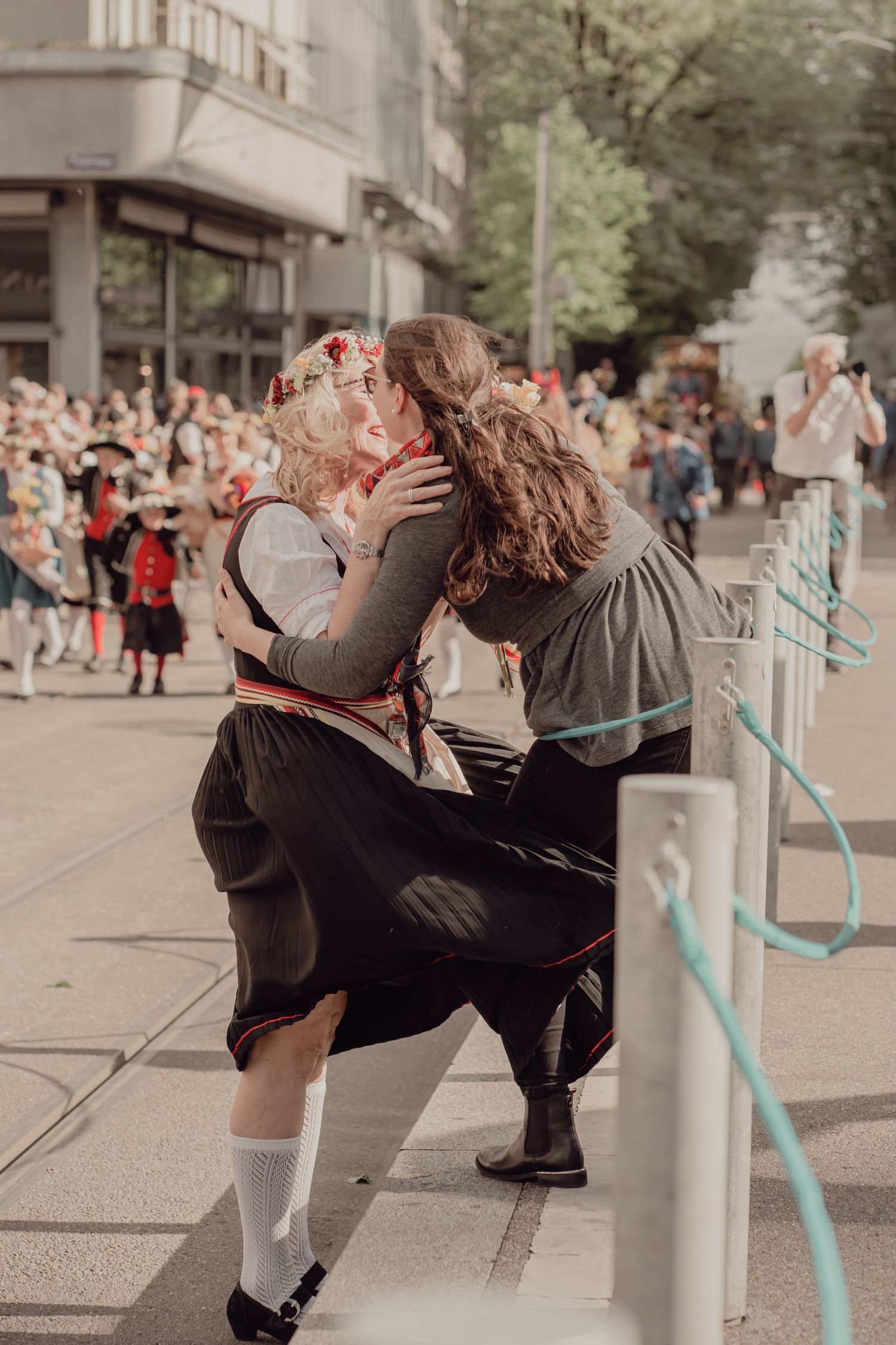 Two women in traditional Swiss folk costumes sharing a warm embrace while watching a folk parade on a sunny city street.