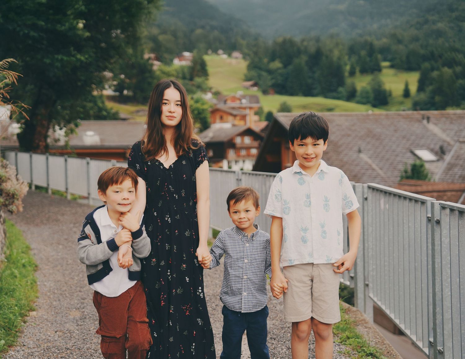 A teenage girl holds hands with three young boys on a scenic Swiss mountain village path.