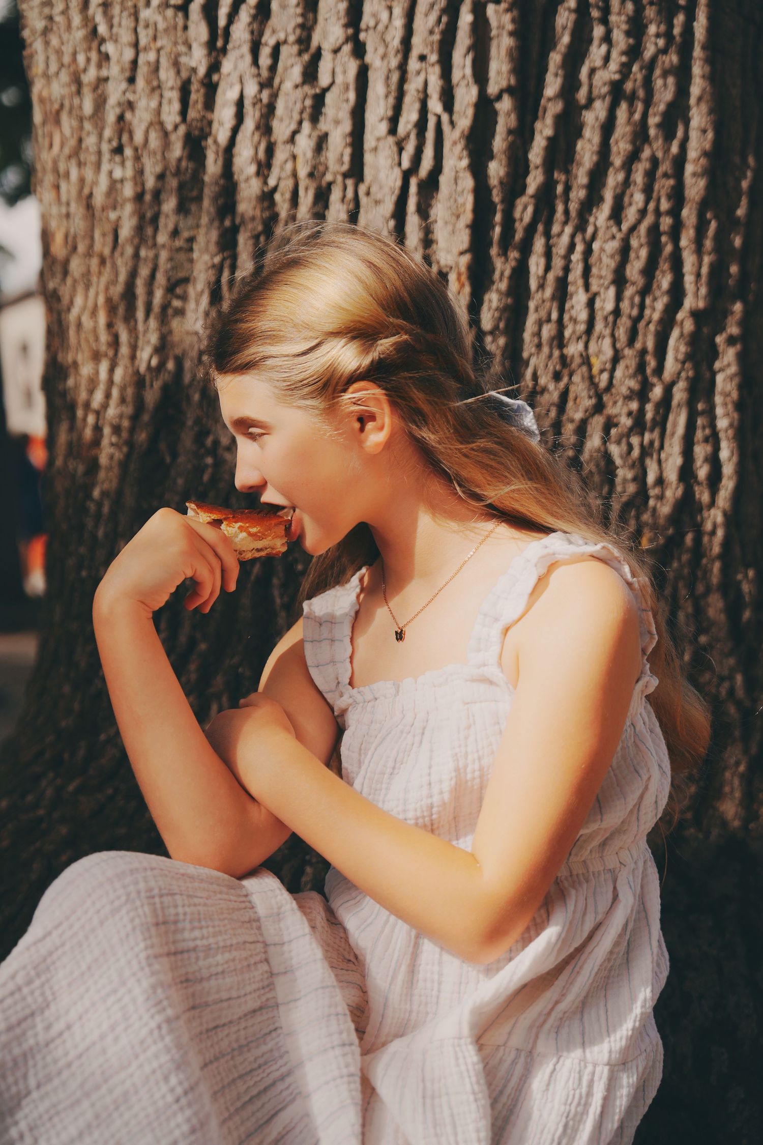 Young blonde girl in white dress eating a snack while sitting against a tree.