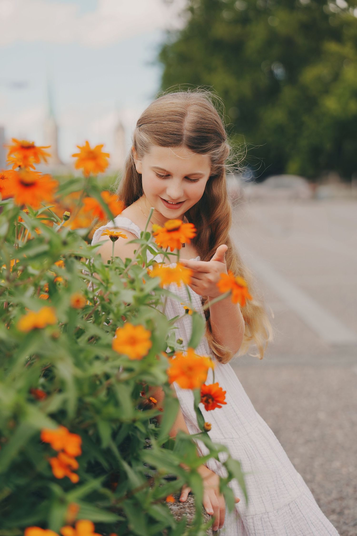 Smiling girl in white dress touching bright orange flowers in urban garden setting.