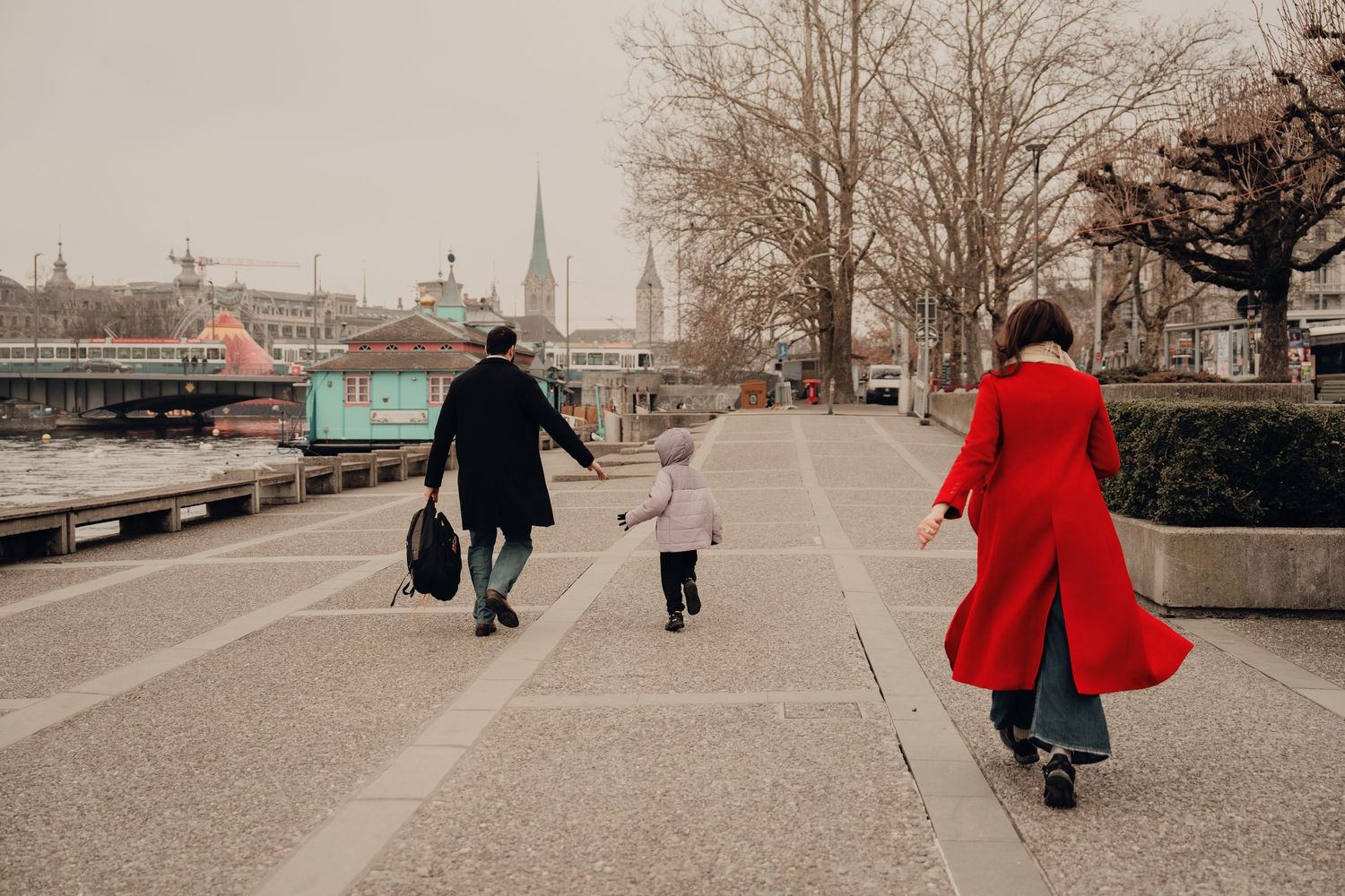 Family of three walking along a riverside promenade, man in black coat holding child's hand with woman in red.