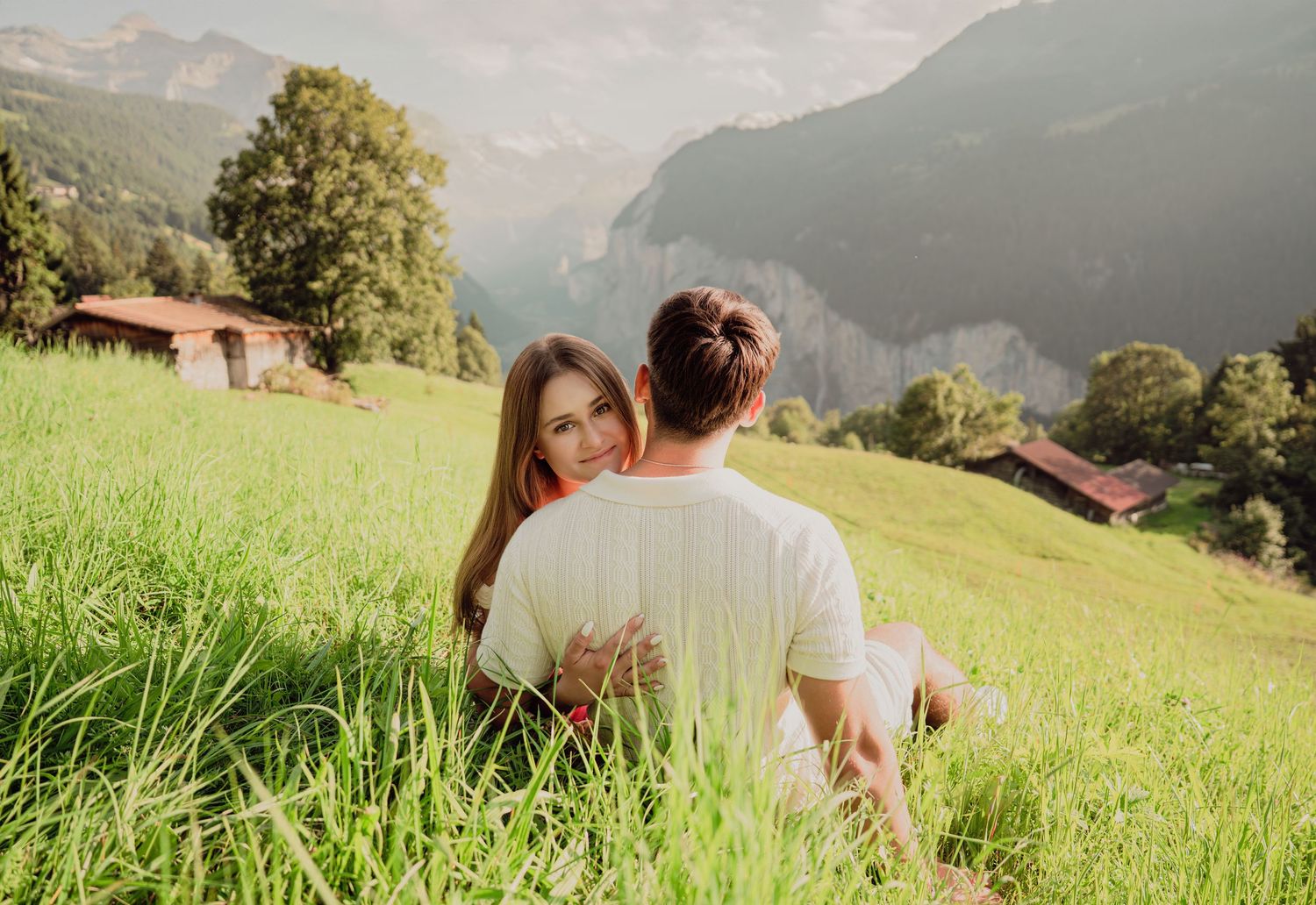 A couple sits embracing on a green hillside with stunning alpine mountain views behind them.