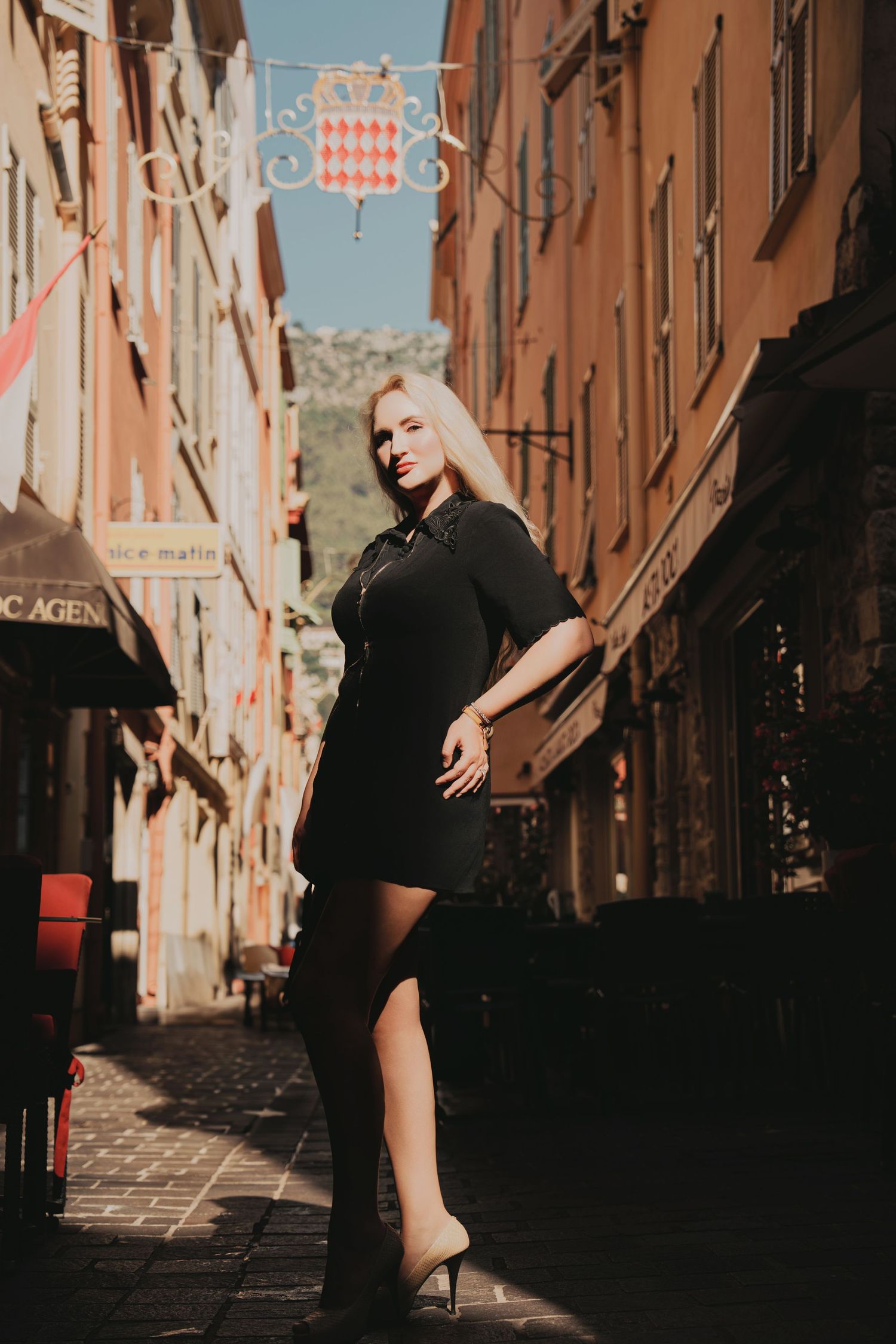 Blonde woman in black mini dress posing in narrow European cobblestone street alley.