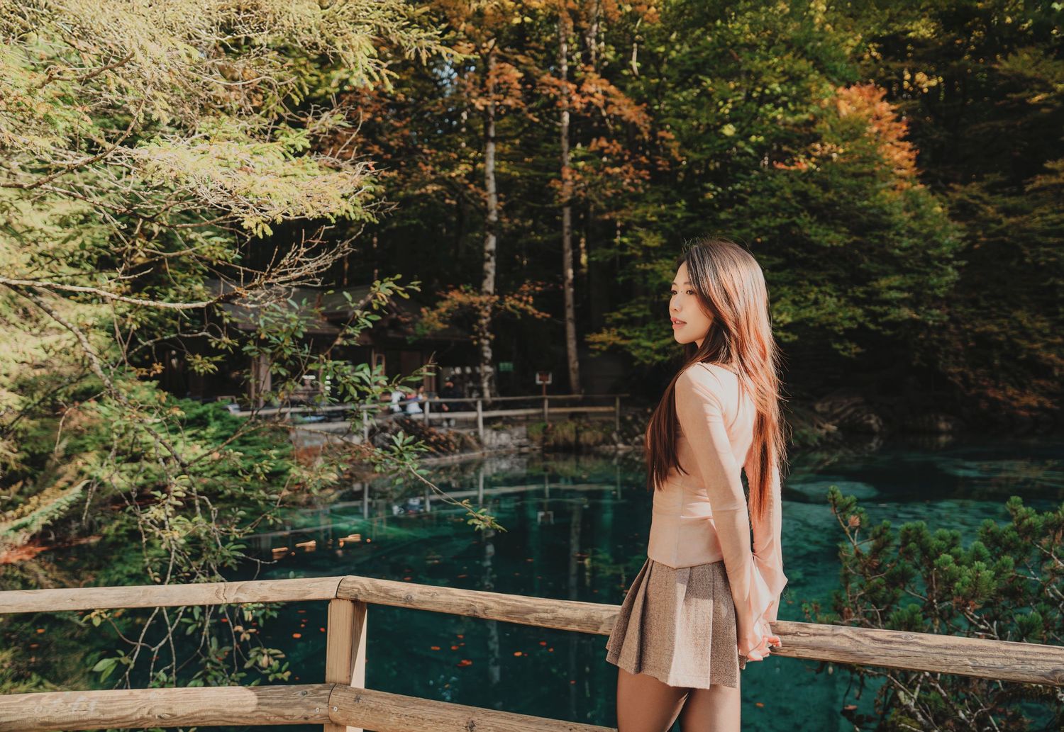 Young woman in beige outfit stands by wooden fence overlooking a vivid turquoise lake in autumn forest.