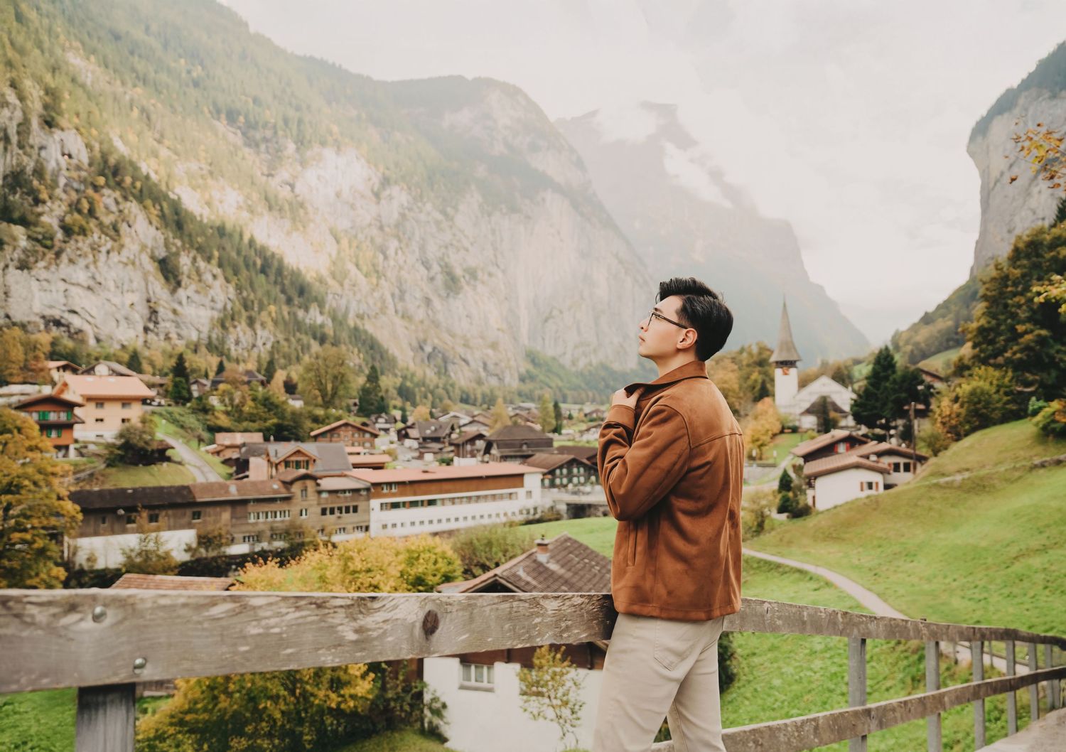 Man in brown jacket standing by a wooden fence admiring a scenic Swiss mountain village landscape.