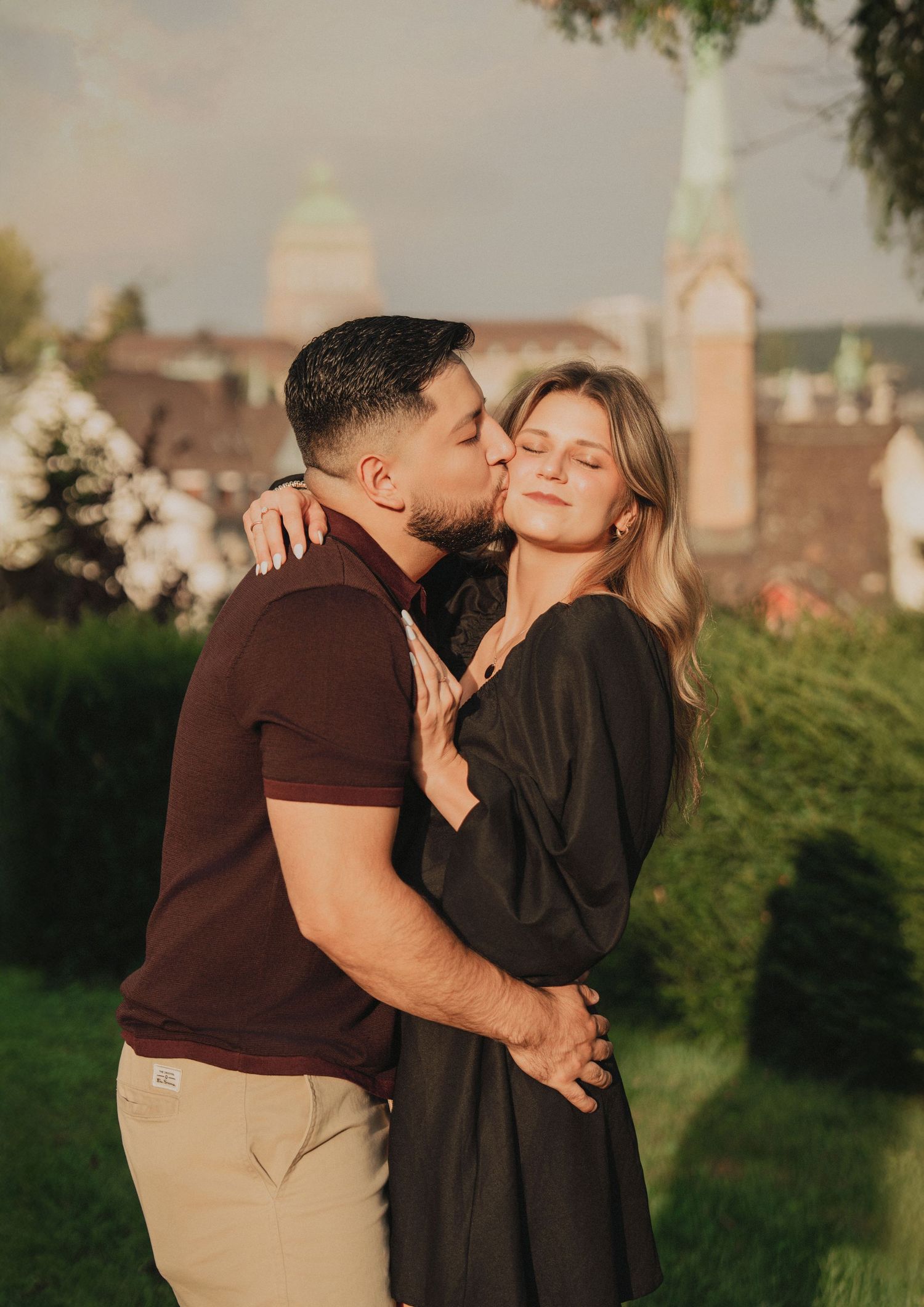 Man tenderly kissing woman on the cheek while embracing in a lush garden with Zurich skyline behind.