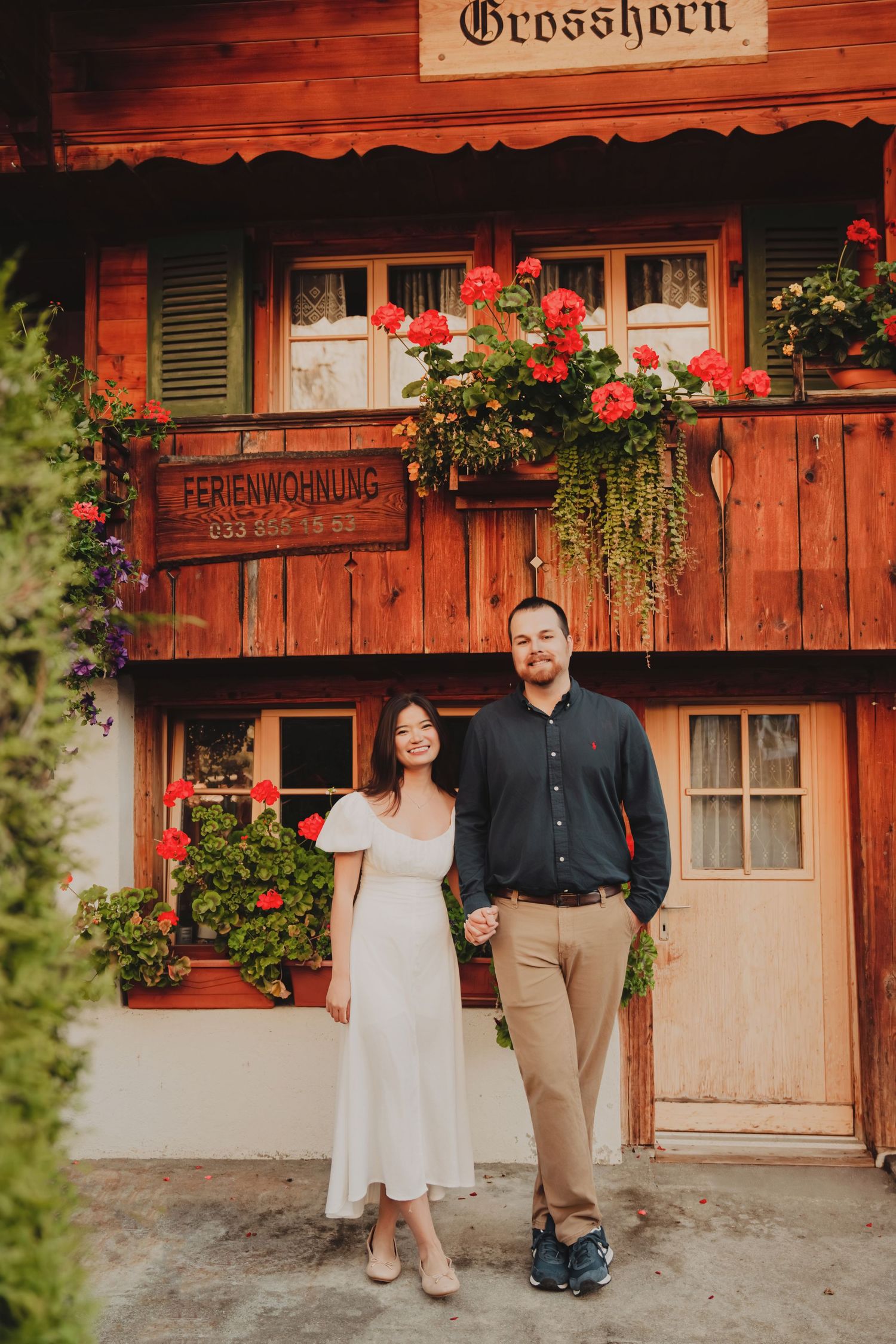 Couple posing hand in hand before the iconic Grosshorn Ferienwohnung in a Swiss mountain village.