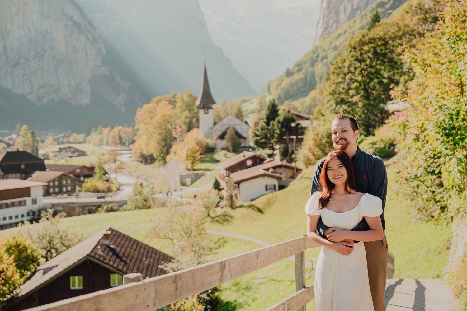 A happy couple hugging on a balcony overlooking a picturesque Swiss village nestled in a mountain valley.