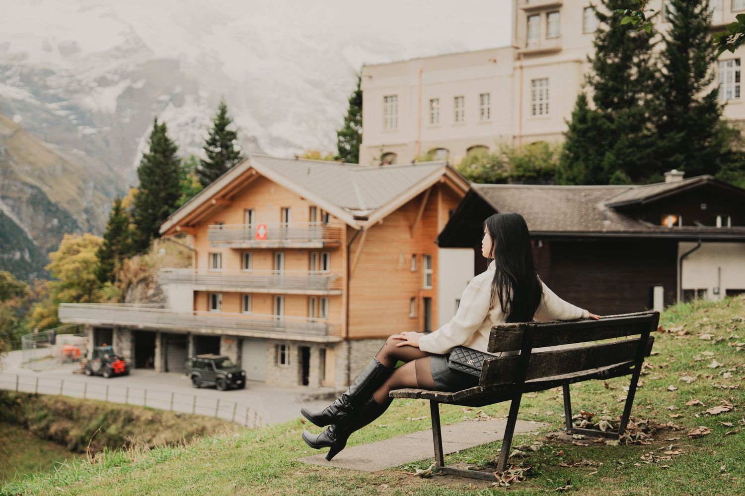 Asian woman in white sweater sits on bench looking down at phone with Swiss mountain village behind her.