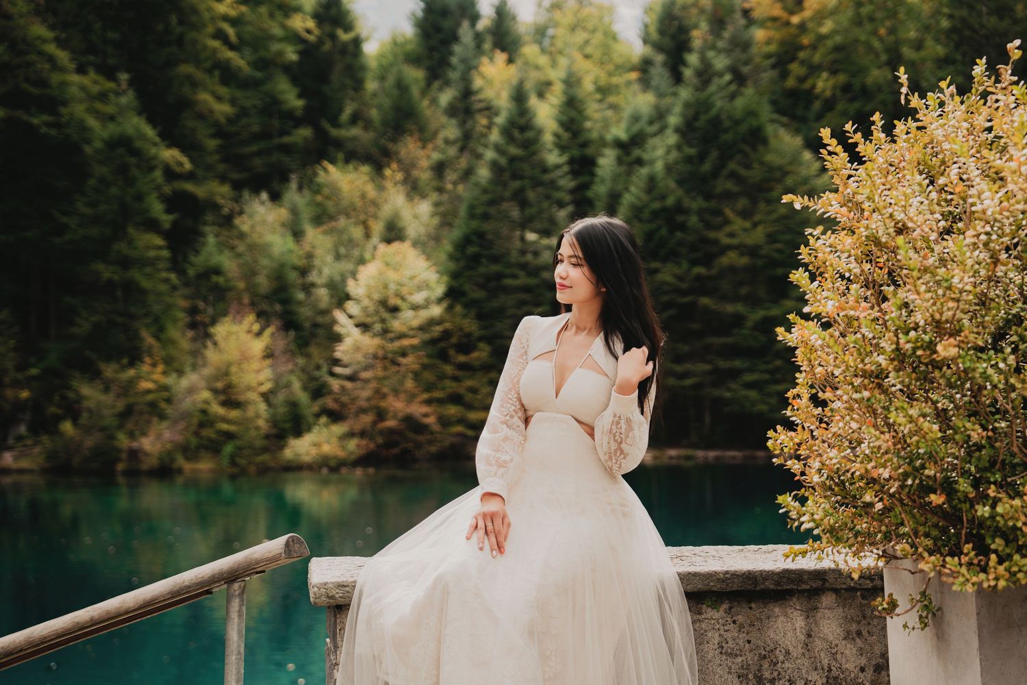 Woman in white dress poses by turquoise lake with autumn forest backdrop.
