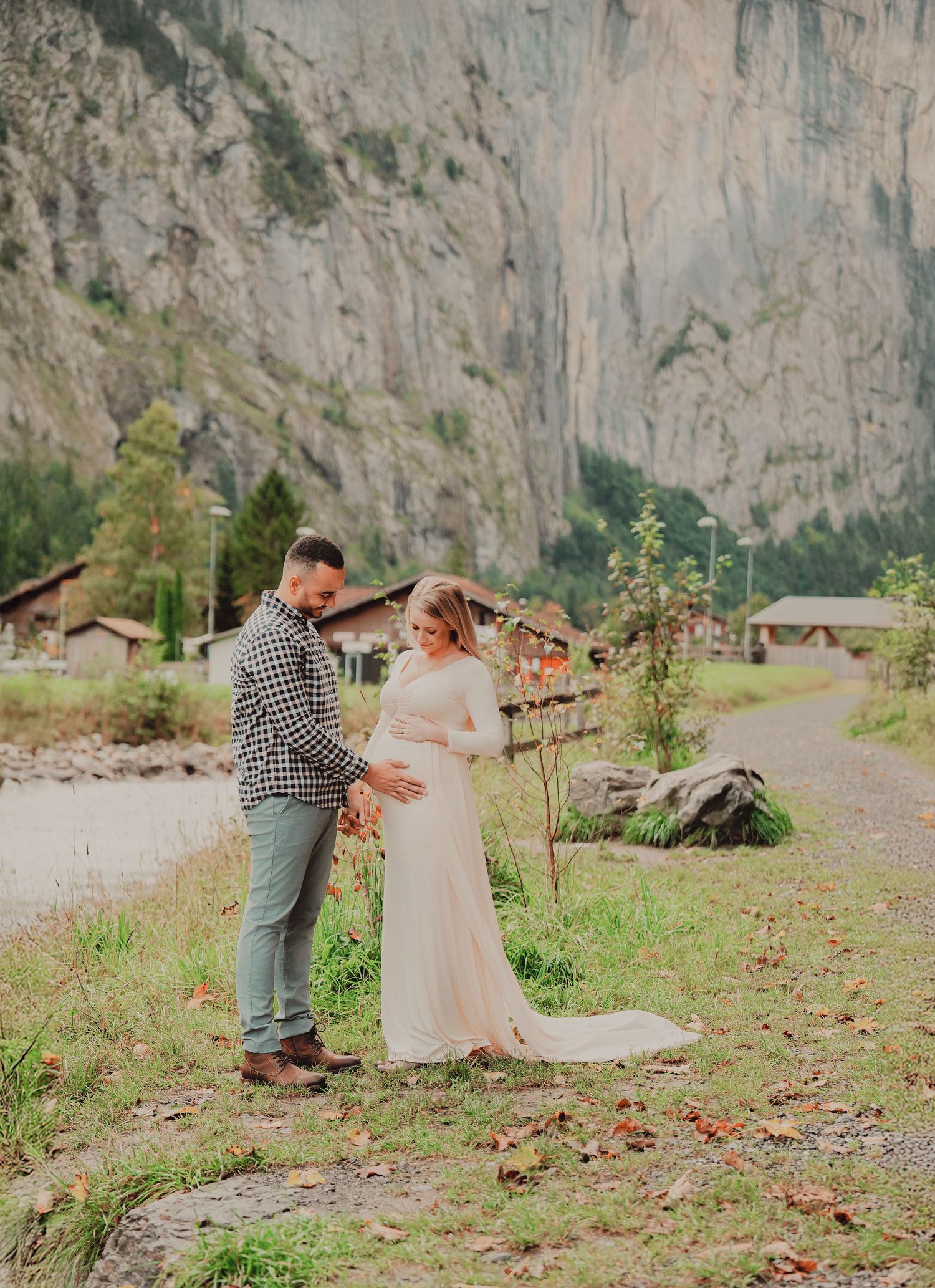 Couple tenderly touching pregnant belly during maternity photoshoot in scenic Swiss alpine valley with dramatic cliffs.