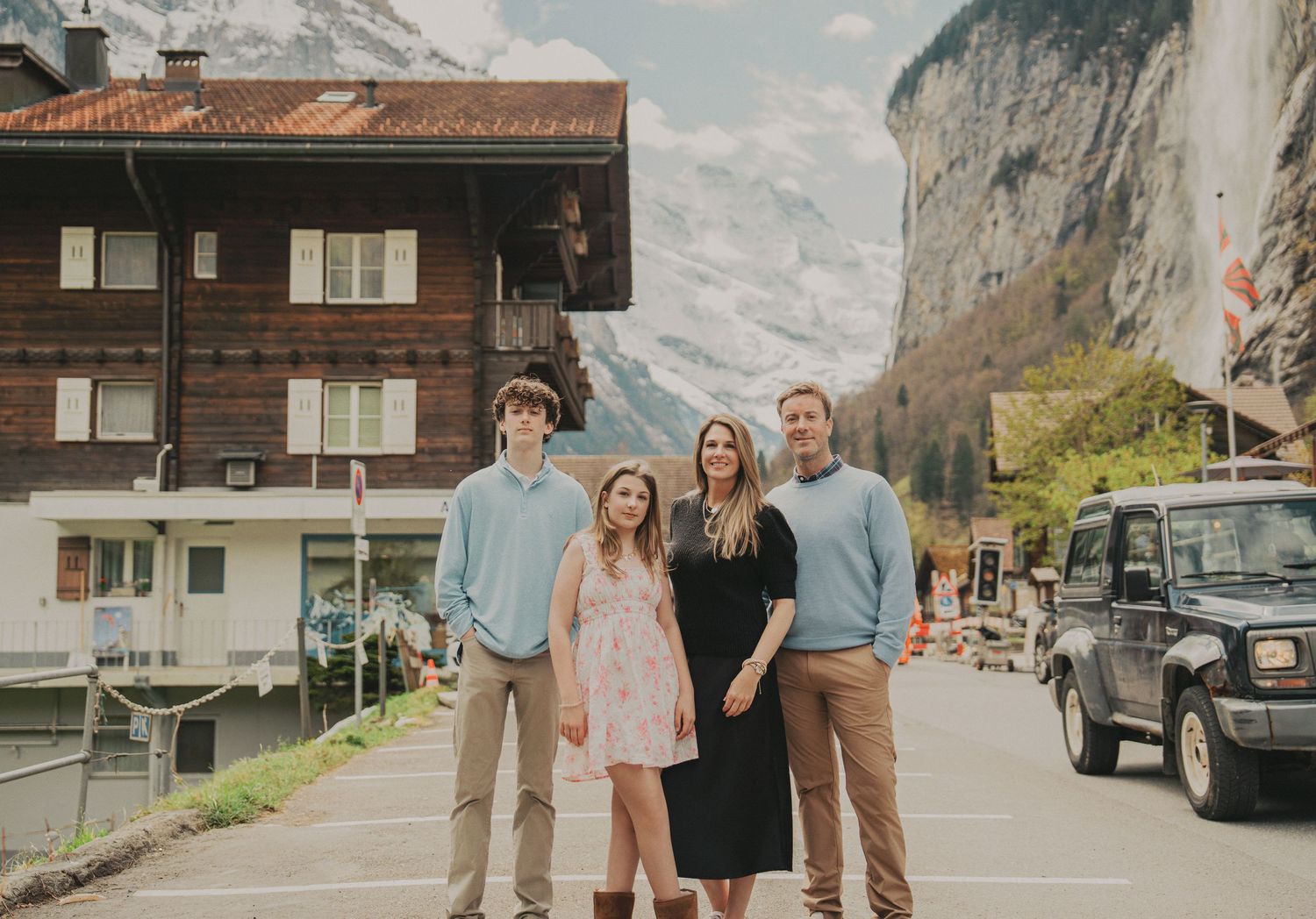 A family of four smiles together on a village street with Swiss alpine chalets and mountains behind them.