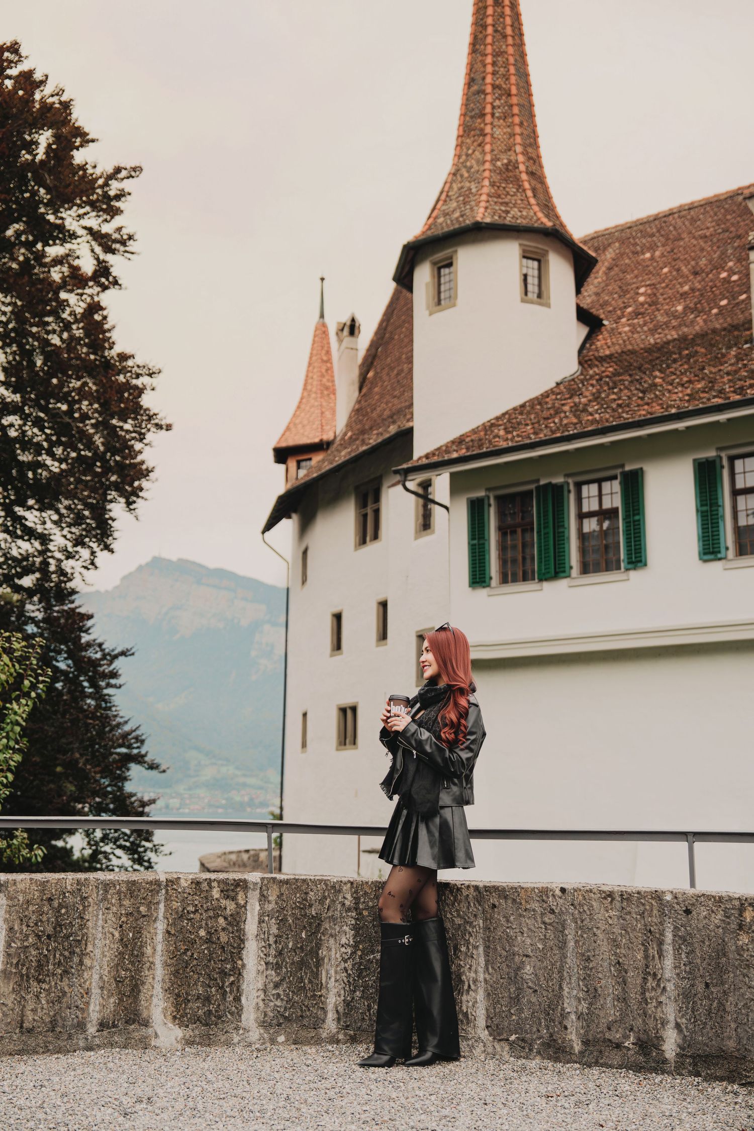 Red-haired woman in black leather outfit stands before a white Swiss castle with terracotta spire on overcast day.