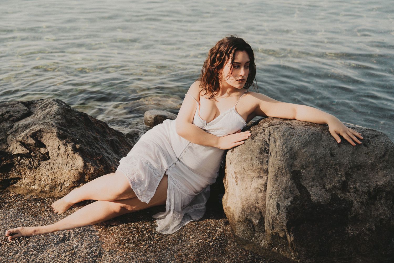 Woman in white dress posing on rocks by the ocean shore.