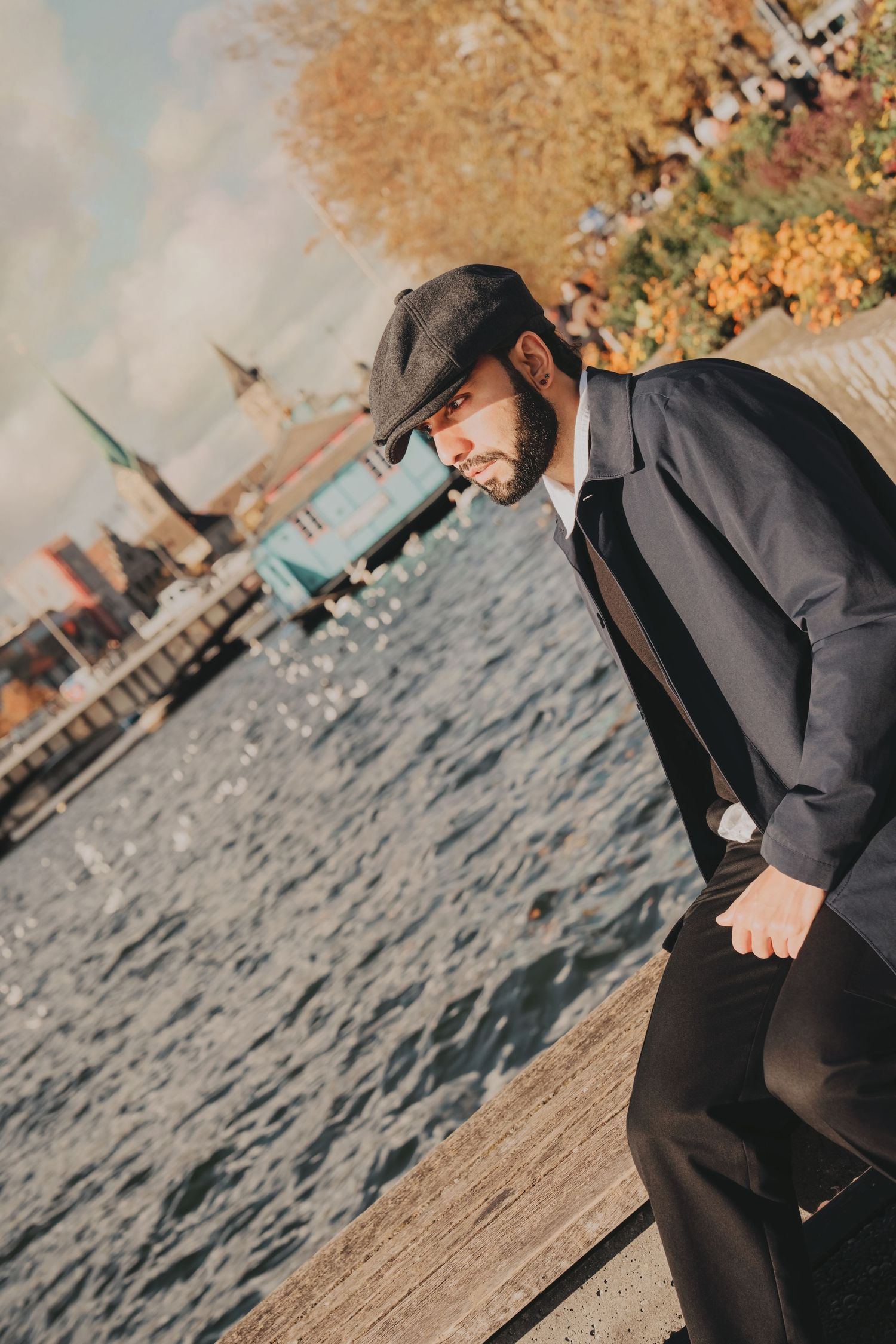 Stylish bearded man in dark suit and flat cap leans forward by a sunlit riverside in autumn.