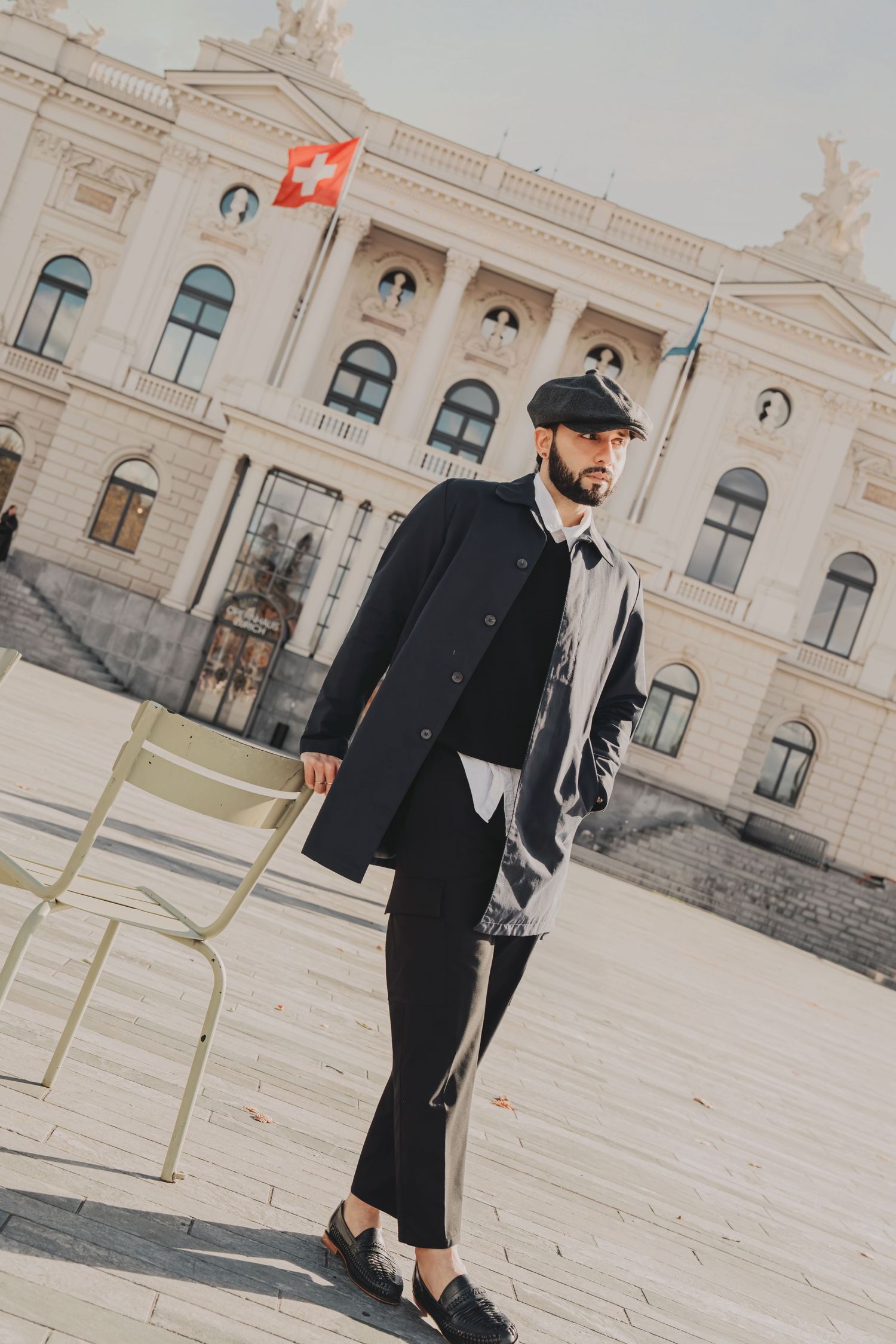 Stylish bearded man in black coat and flat cap posing outside a grand European building with Swiss flag.