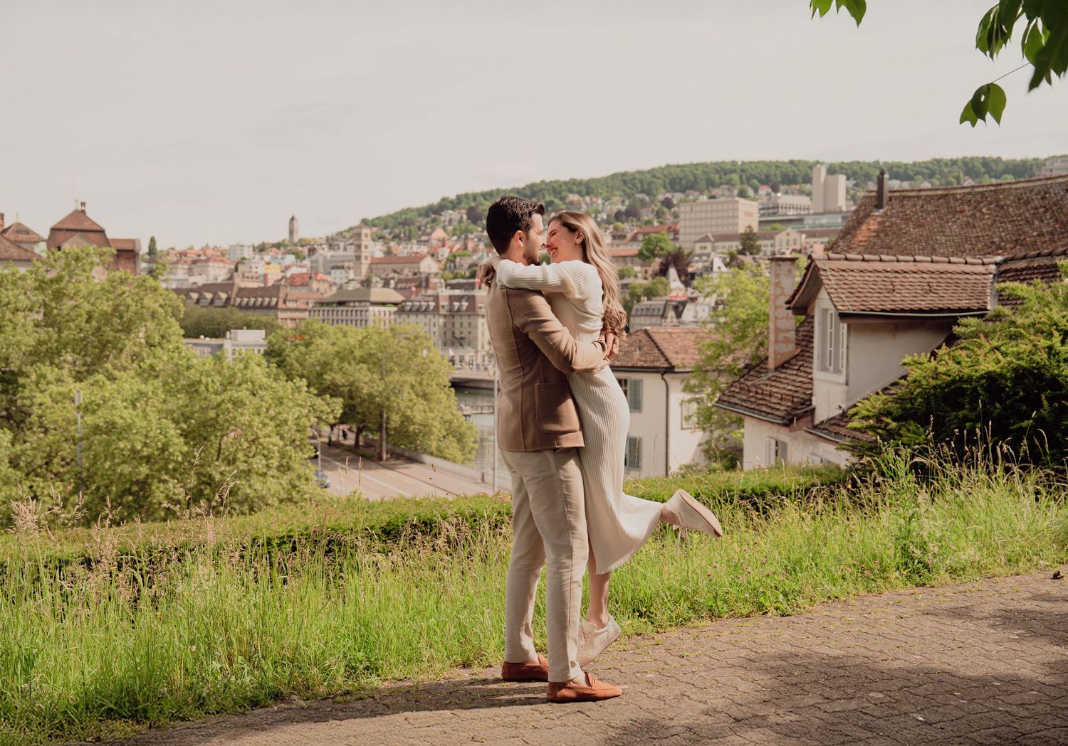 Couple embracing and kissing on scenic overlook with European city rooftops and green hills behind.