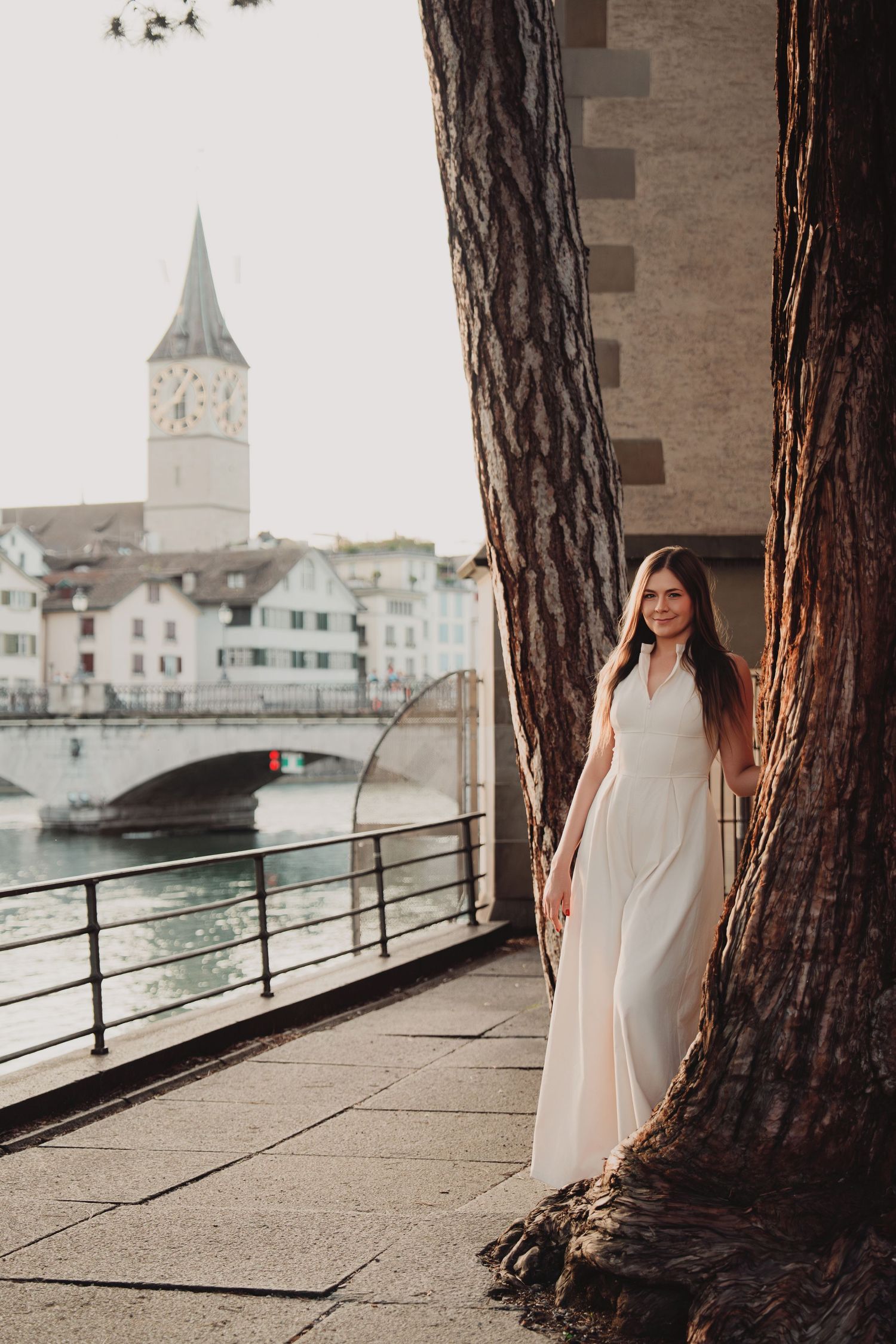 Woman in white dress standing between large tree trunks by Zurich river with St. Peter church tower.