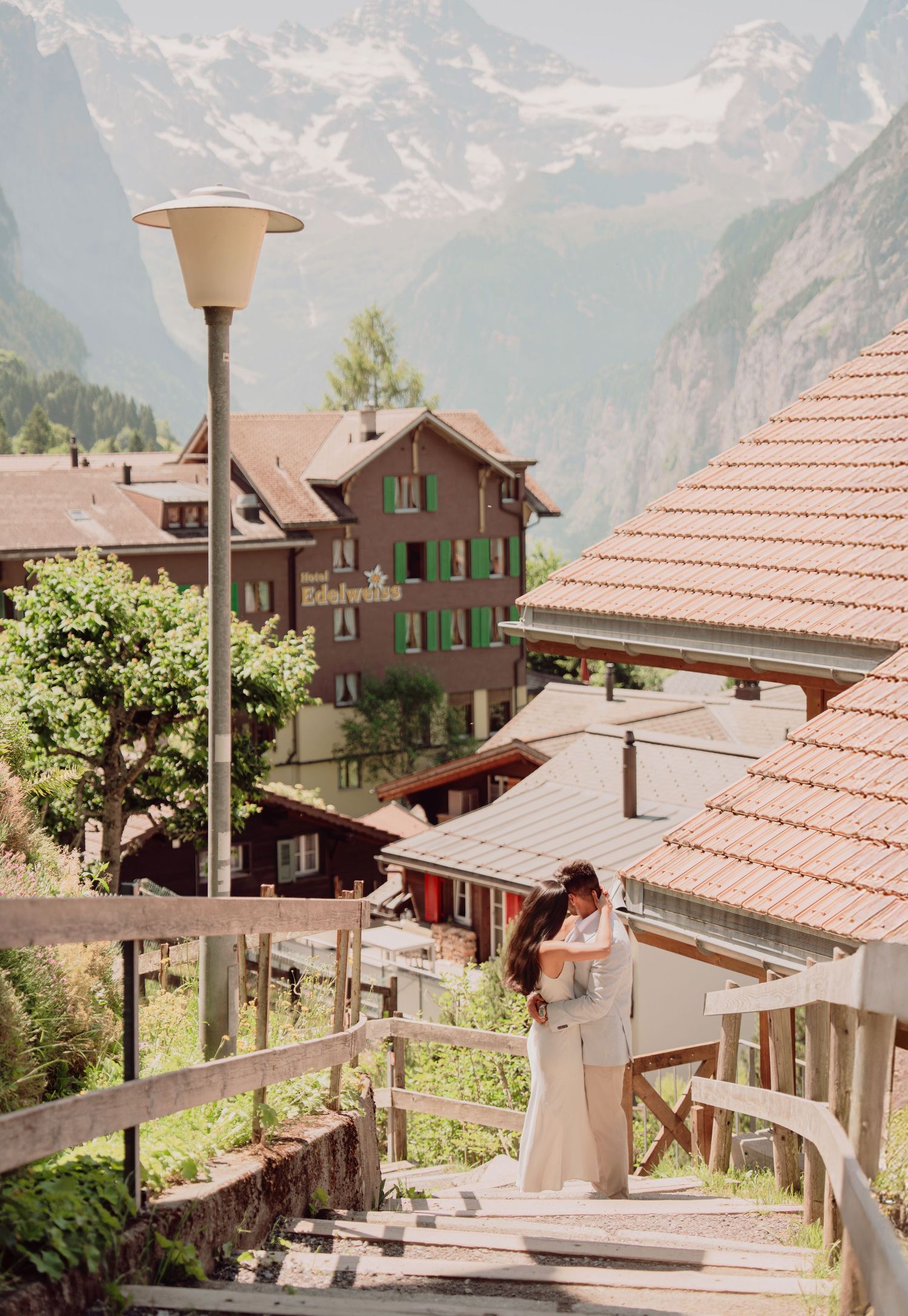 Couple embracing on wooden stairs in a picturesque Swiss village with traditional chalets and mountain views behind them.