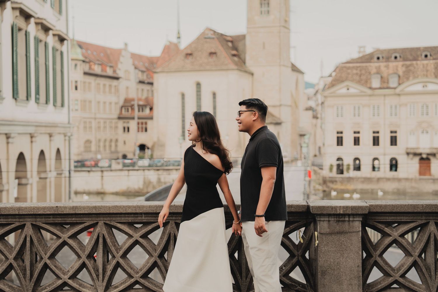 Stylish couple holding hands on a Zurich bridge, posing back-to-back with historic buildings behind.