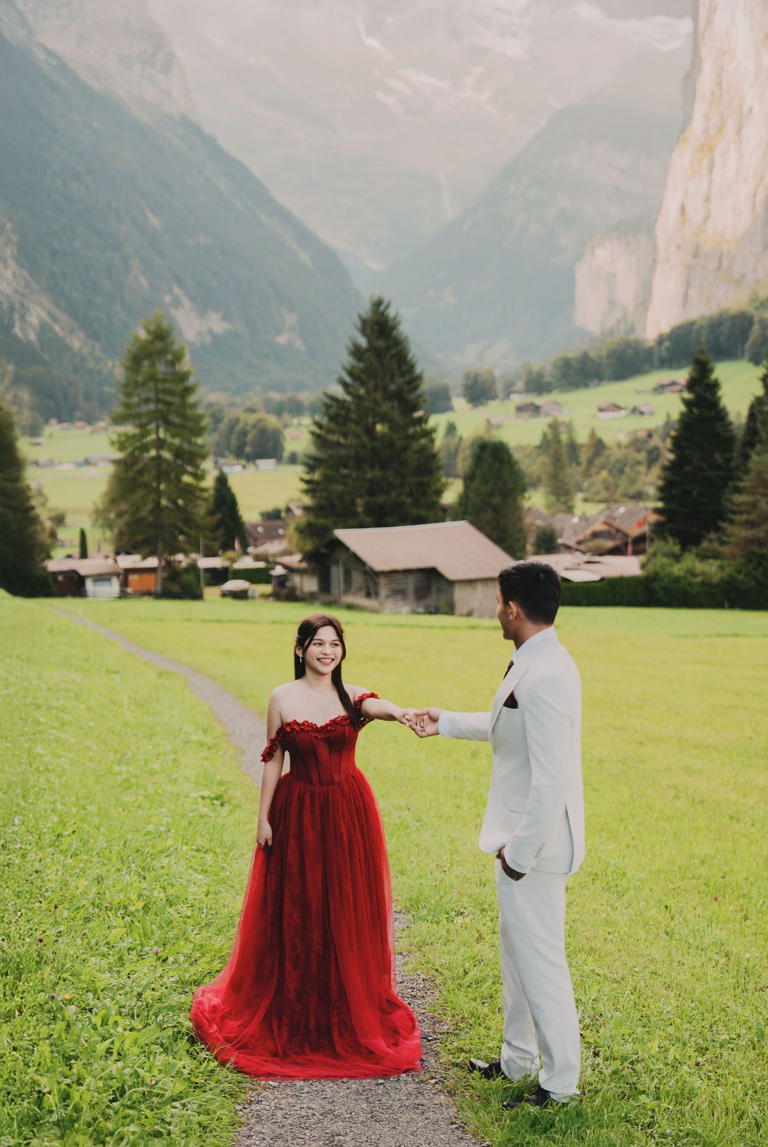 Bride in red tulle gown holds groom's hand on scenic Swiss alpine meadow path.